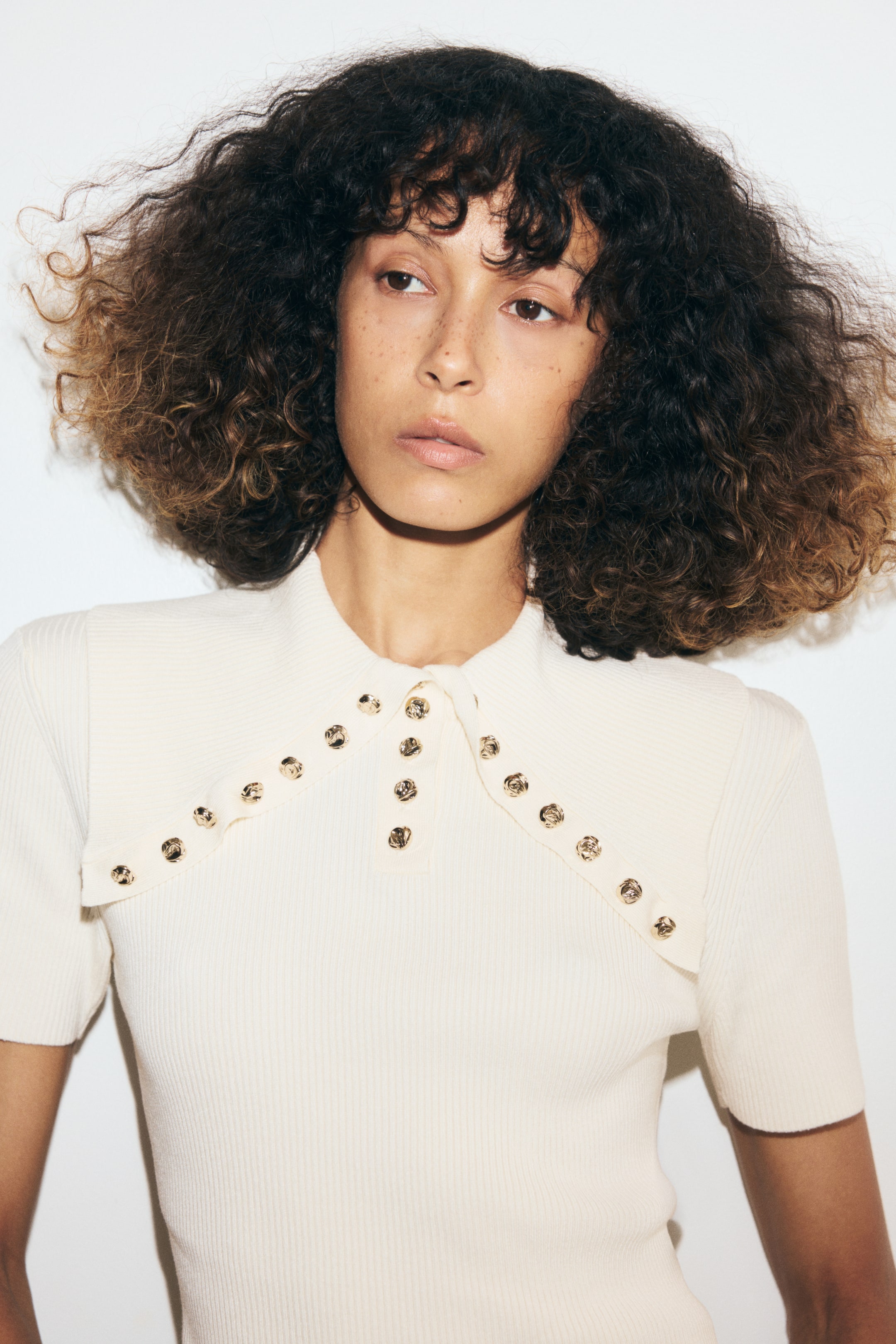 View larger image: The model is wearing a cream-colored, fitted rib-knit top featuring short sleeves, a central button placket, and a large collar accented with decorative gold buttons.