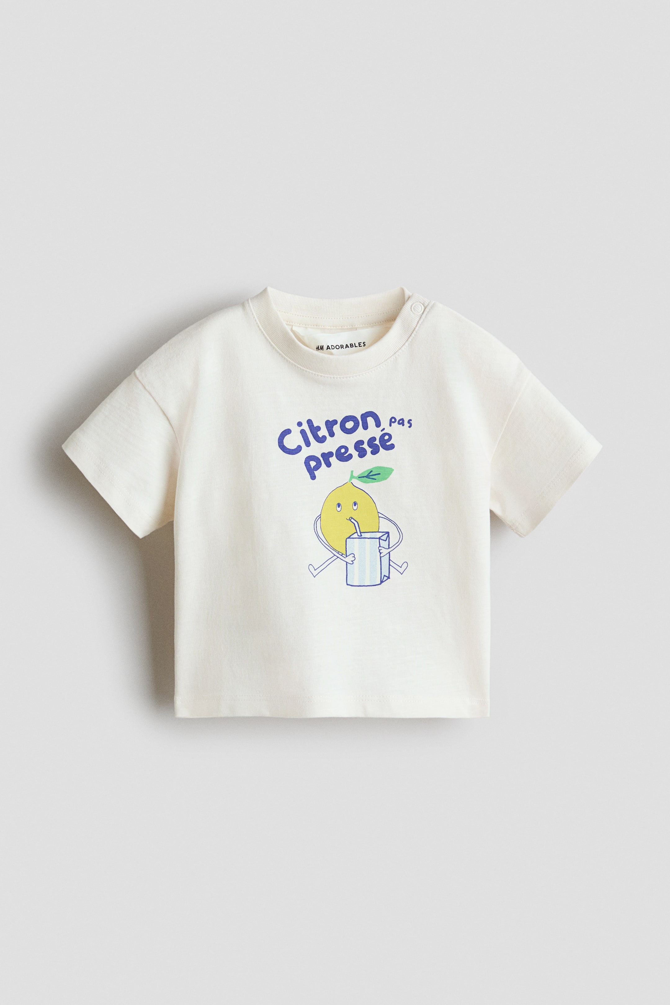 Agrandir l'image: Un t-shirt blanc cassé est présenté de face, avec un col rond bordé de côtes et deux pressions blanches sur l'épaule gauche, ainsi que des manches courtes à épaules tombantes. Un motif brodé coloré est visible à l'avant, montrant un personnage de citron en dessin animé buvant d'une boîte à jus rayée, avec le texte bleu "Citron pas pressé" au-dessus.