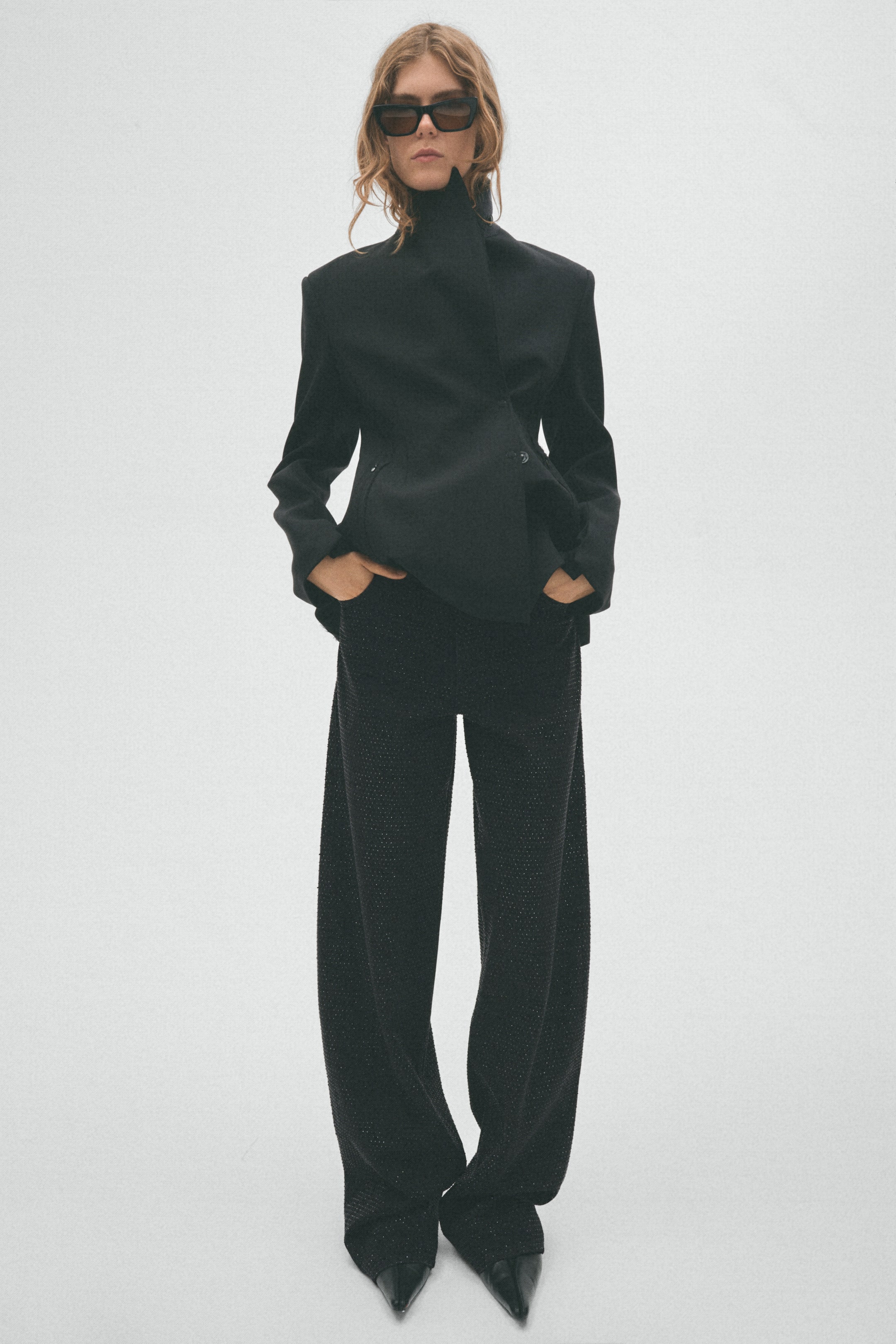 View larger image: A black blazer featuring a high stand collar, structured shoulders, and an asymmetric front closure with one visible button is worn by the model. The model styles the blazer with black wide-leg trousers and black pointed-toe shoes.