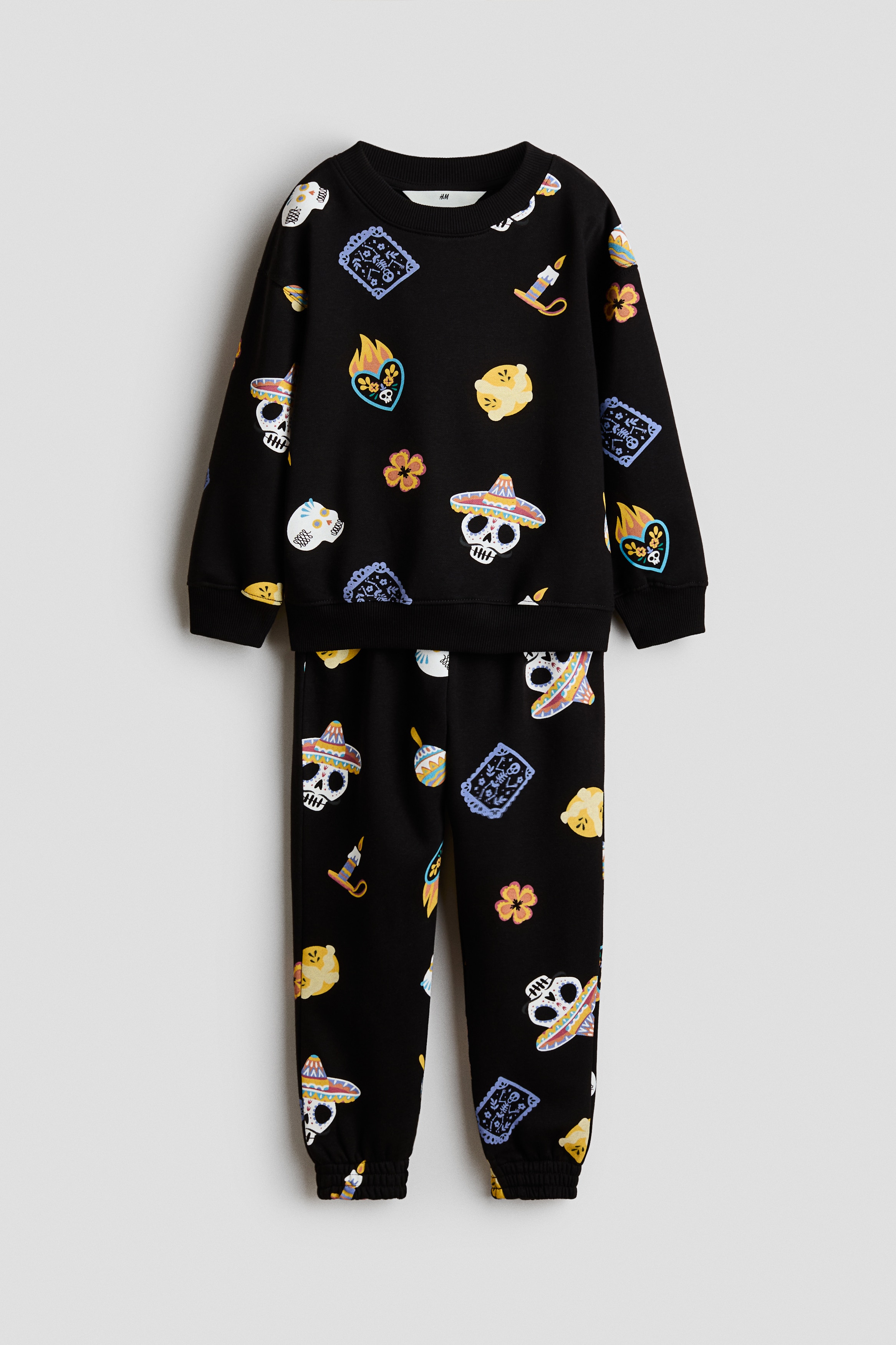 A black matching sweater and track pants set is shown from the front, featuring an all-over colorful print of cartoon skulls, flowers, and festive icons. The long-sleeved sweater is designed with a round ribbed neckline, dropped shoulders, and ribbed cuffs and hem, while the matching track pants feature an elasticated waistband and elasticated hems.