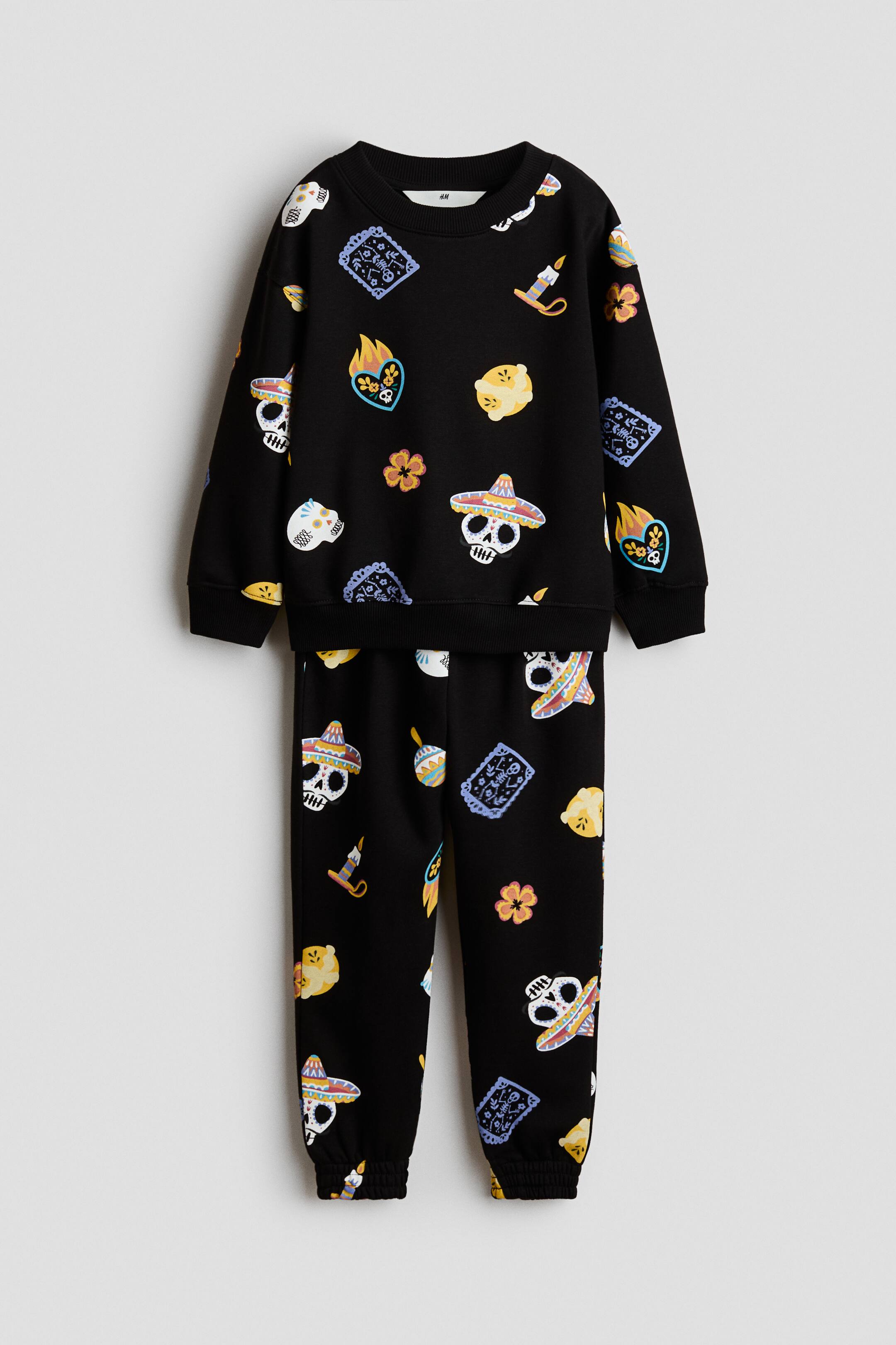 View larger image: A black two-piece loungewear set is displayed, featuring an allover colorful pattern of stylized skull motifs wearing sombreros, candles, flaming hearts, flowers, and rectangular patterned shapes. The crew-neck sweatshirt features long sleeves with ribbed cuffs and a ribbed hem, and the matching jogger pants feature ribbed ankle cuffs.