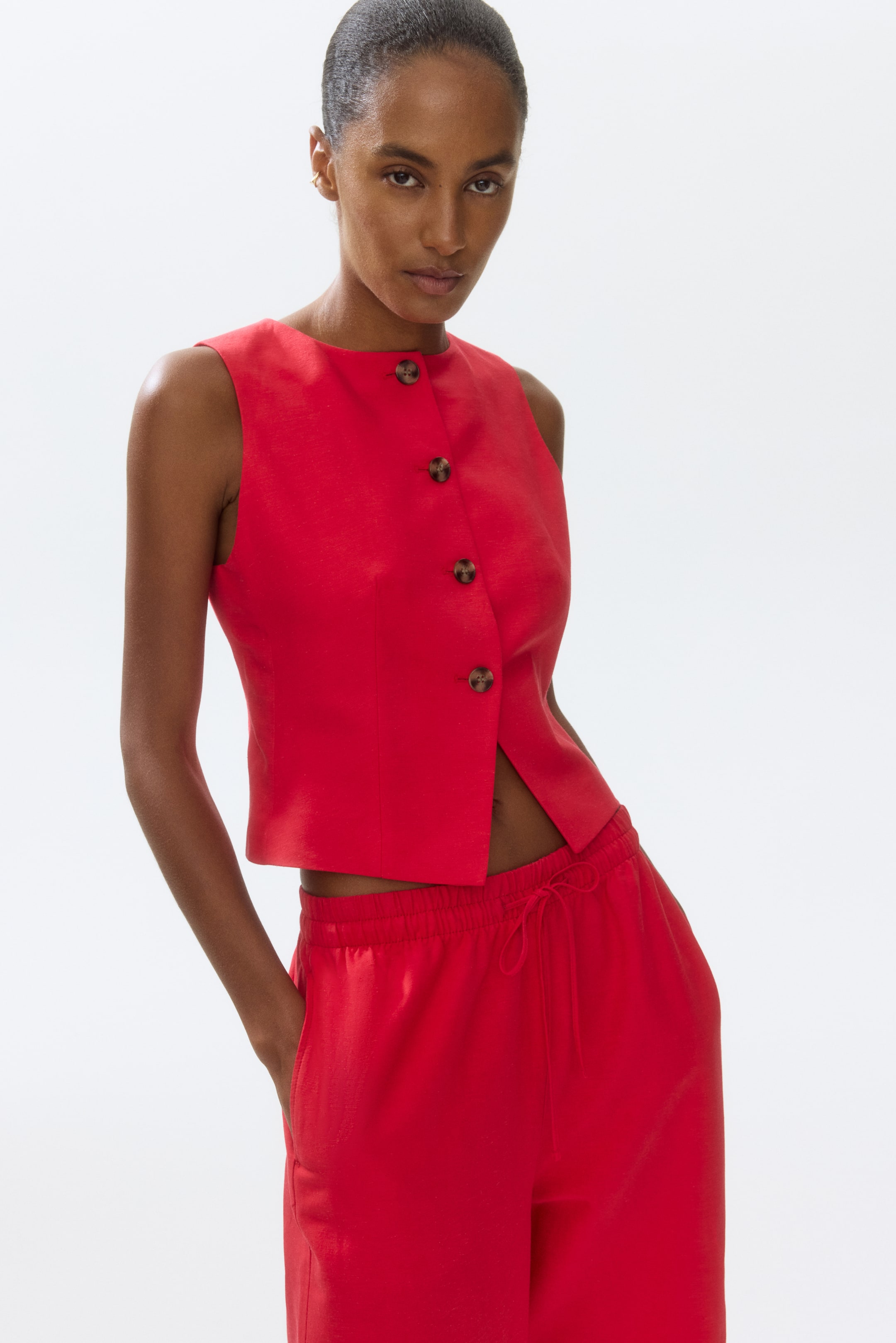 View larger image: The model is donning a red tailored waistcoat crafted from woven fabric. It features a round neckline, with brown buttons running down the front to the waist. Notably, the lowest button is left undone.