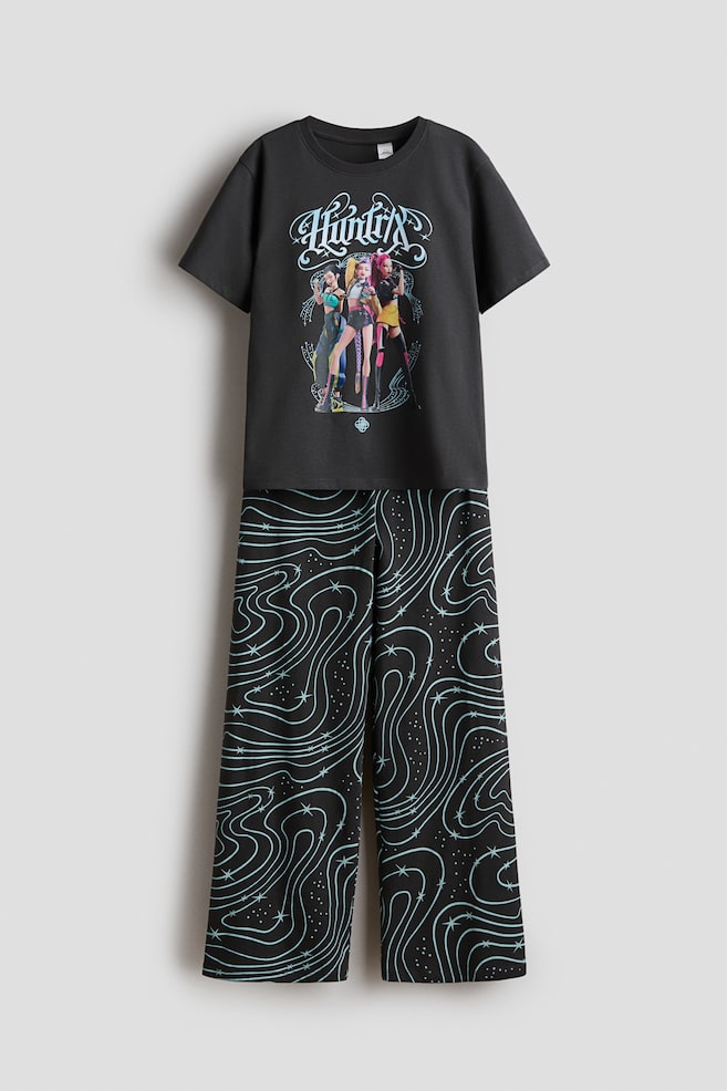 Ein zweiteiliger Pyjama wird von vorne gezeigt und besteht aus einem schwarzen T-Shirt mit kurzen Ãrmeln und einer weit geschnittenen Hose. Das T-Shirt zeigt einen Grafikprint mit drei animierten Figuren unter stilisiertem Schriftzug im Gothic-Stil, wÃ¤hrend die schwarze Hose ein hellblaues Muster aus wirbelnden Linien und kleinen Sternen aufweist. - 1