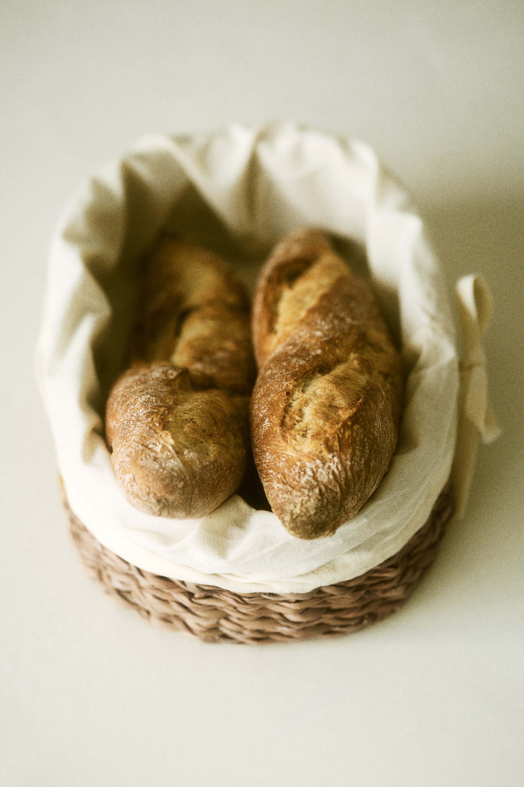 Ingrandisci l'immagine: Due baguette dorate, con incisioni visibili e una leggera spolverata di farina, riposano in un cesto di vimini foderato con un panno color crema.
