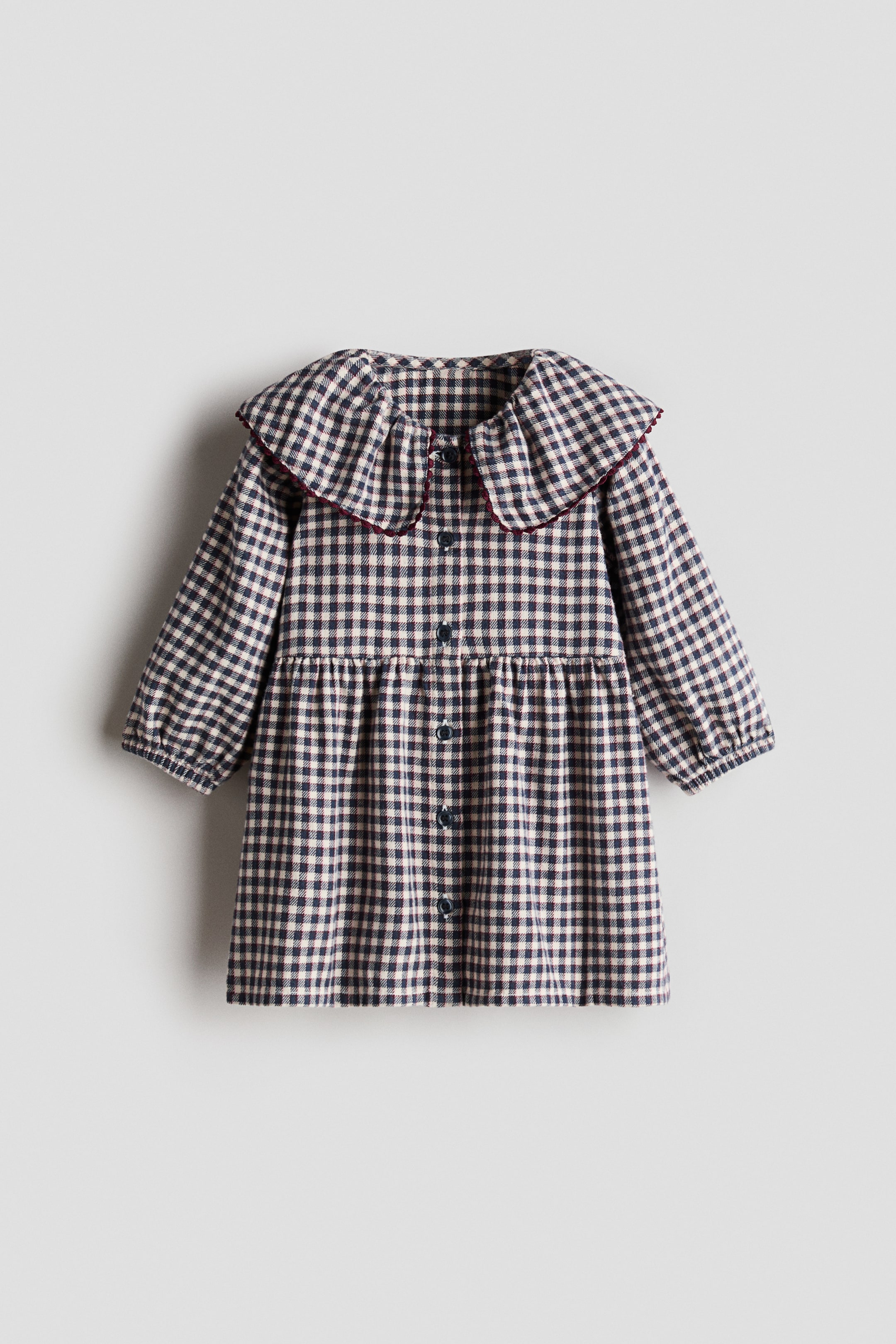 Agrandir l'image: Une robe arbore un motif vichy bleu marine et blanc cassé, avec un grand col à volants bordé de rouge foncé. Les manches longues sont froncées aux poignets, et une patte de boutonnage centrale descend sur le devant, fermée par des boutons foncés, tandis qu’une couture froncée est visible au-dessus de la jupe.