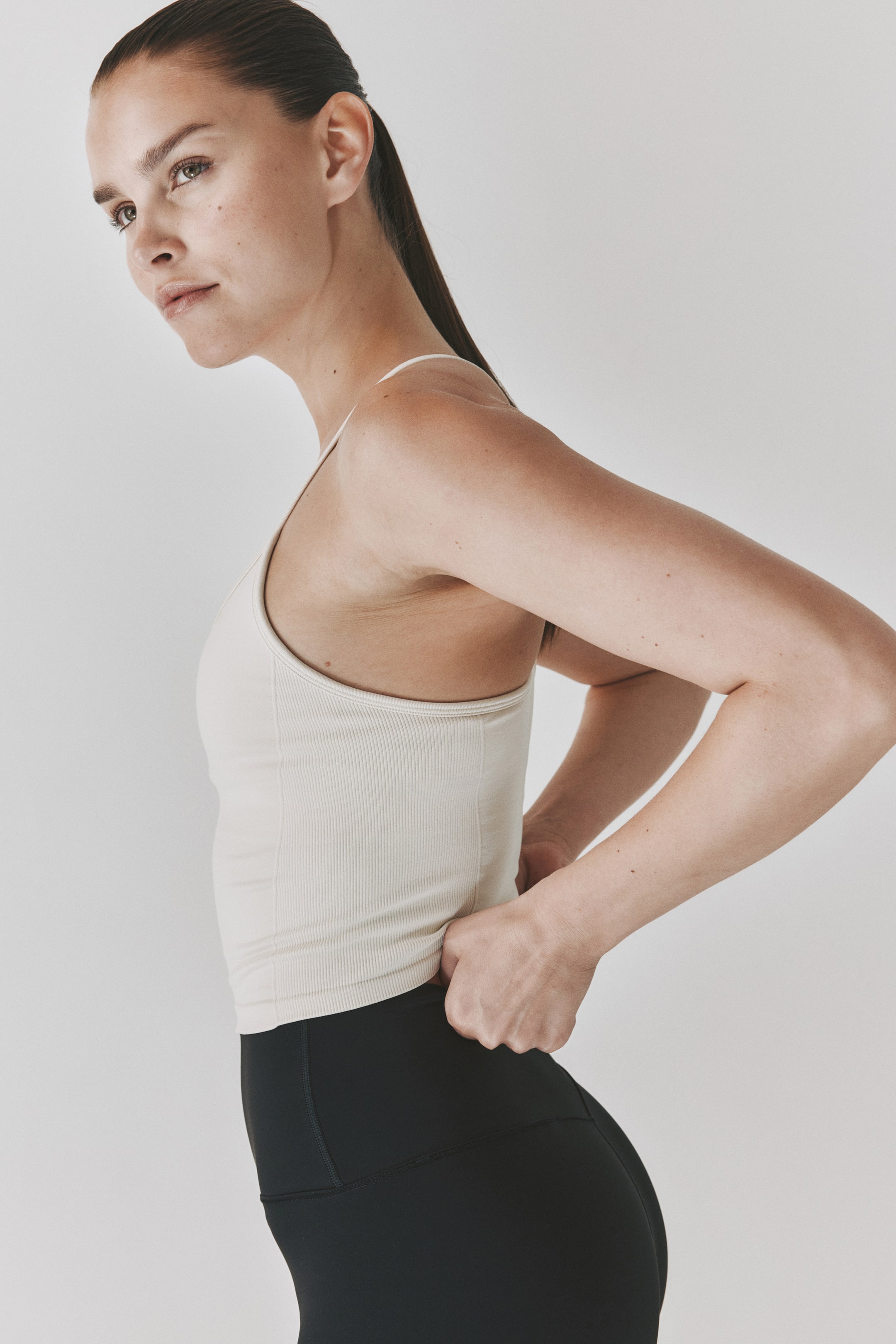 Grotere afbeelding bekijken: Een licht beige geribde tanktop wordt gedragen, met dunne spaghettibandjes en een nauwsluitend zijprofiel. Het wordt gecombineerd met zwarte, nauwsluitende leggings, met naaddetails rond de heup.