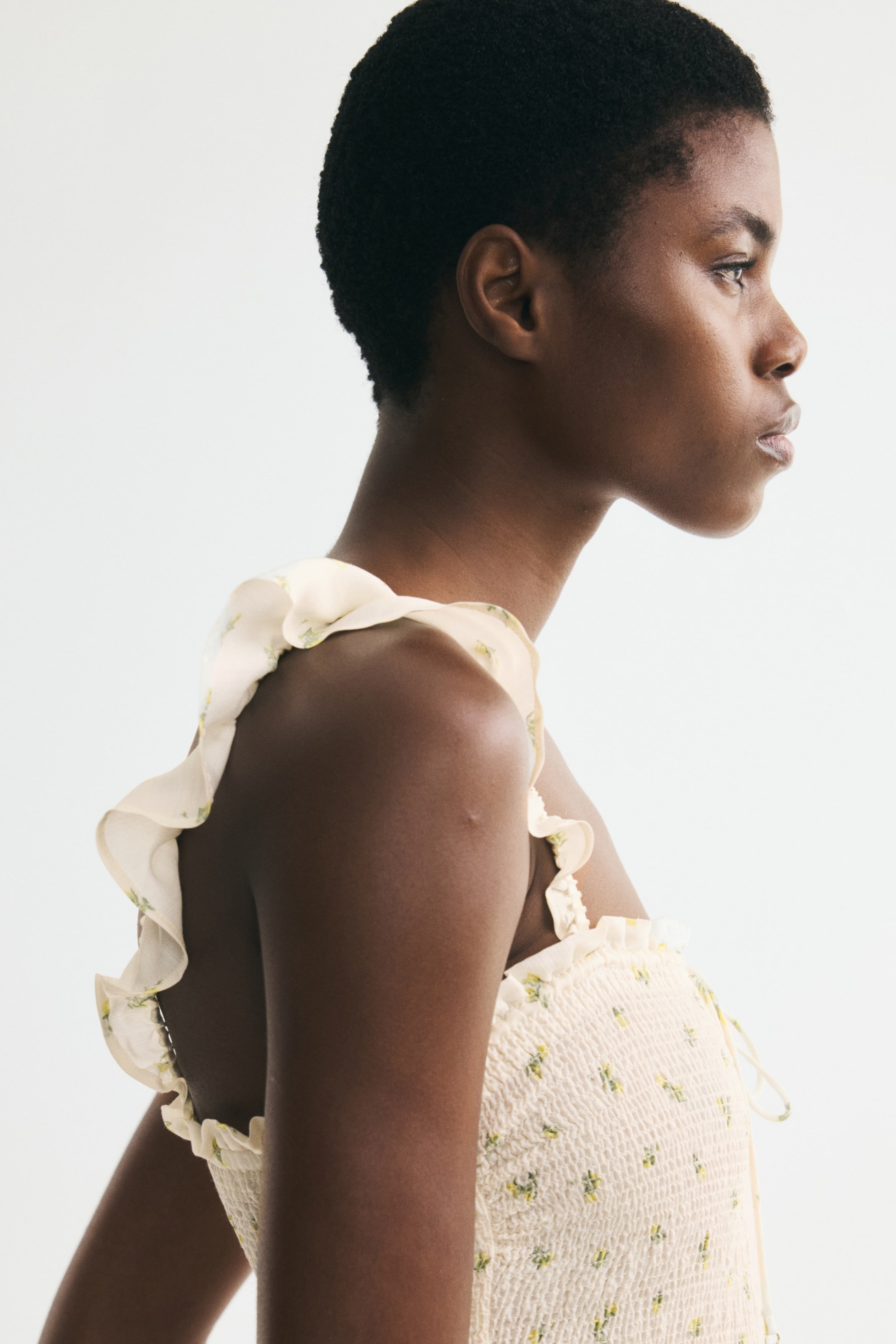 Größeres Bild anzeigen: Ein blassgelbes Kleid mit einem kleinen grünen und gelben Blumenmuster wird vom Model getragen. Es verfügt über mit Rüschen besetzte Schulterträger, ein gesmoktes Oberteil und einen Spaghettizug am Ausschnitt.