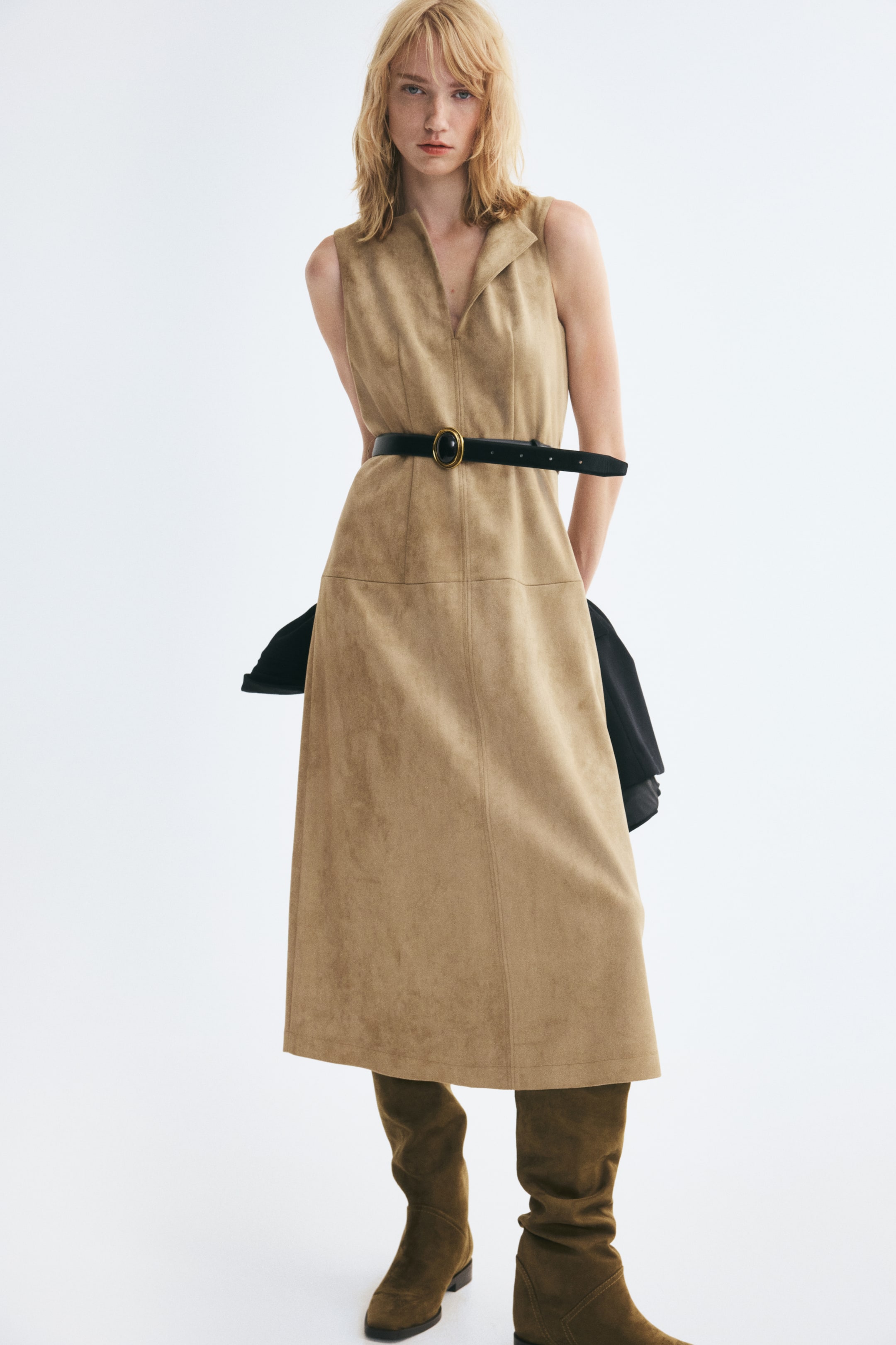 Agrandir l'image: Une robe sans manches beige, longueur mi-mollet, avec une finition douce et légèrement duveteuse, est portée par le mannequin. Elle présente un col rond avec une ouverture en V et une fine ceinture noire cintrée à la taille. La robe est associée à des bottes en daim vert olive montant jusqu’aux genoux.