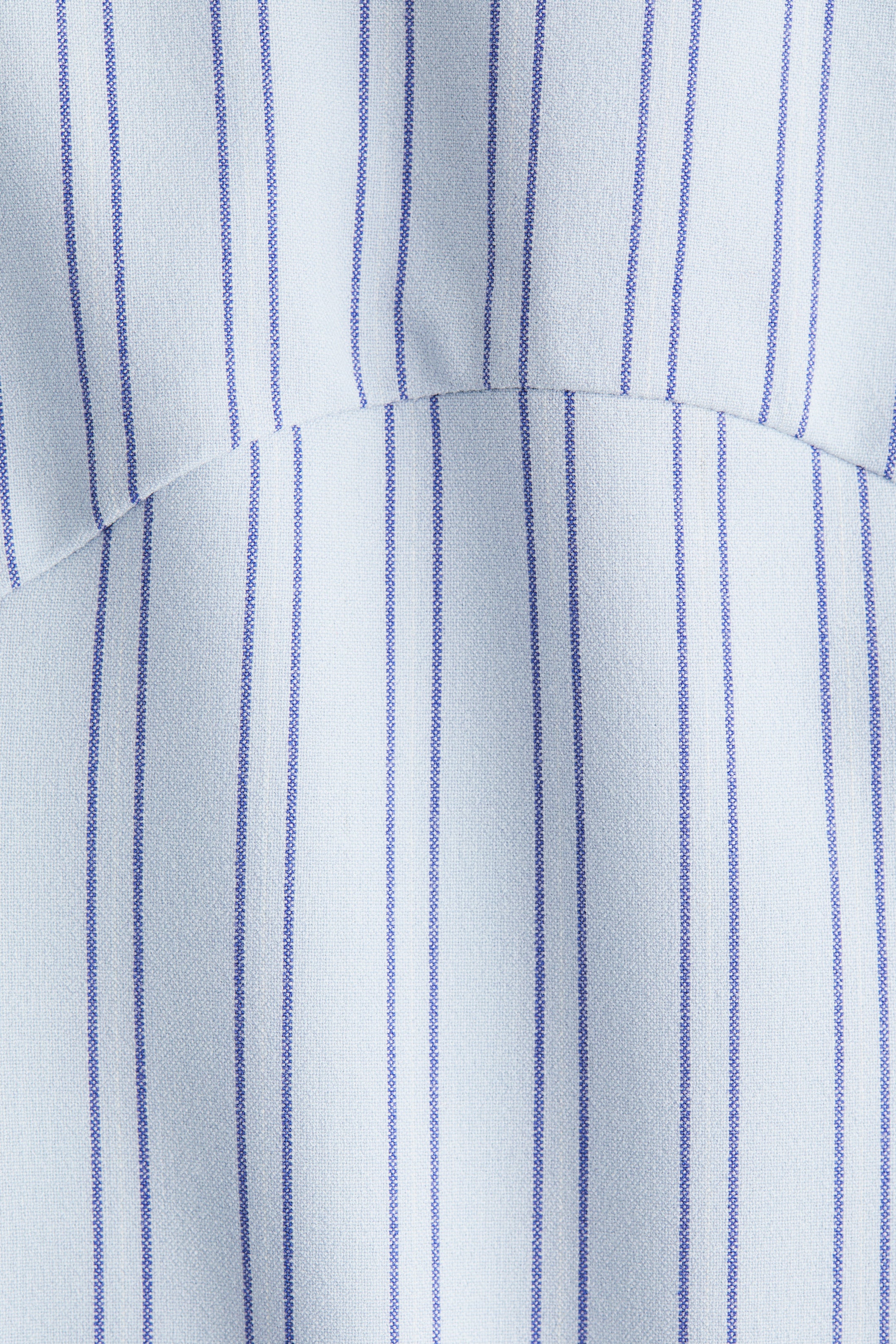 View larger image: The fitted top is featuring a light-colored woven fabric with narrow vertical blue pinstripes. A curved horizontal seam is visible across the fabric, contributing to its contoured shape.