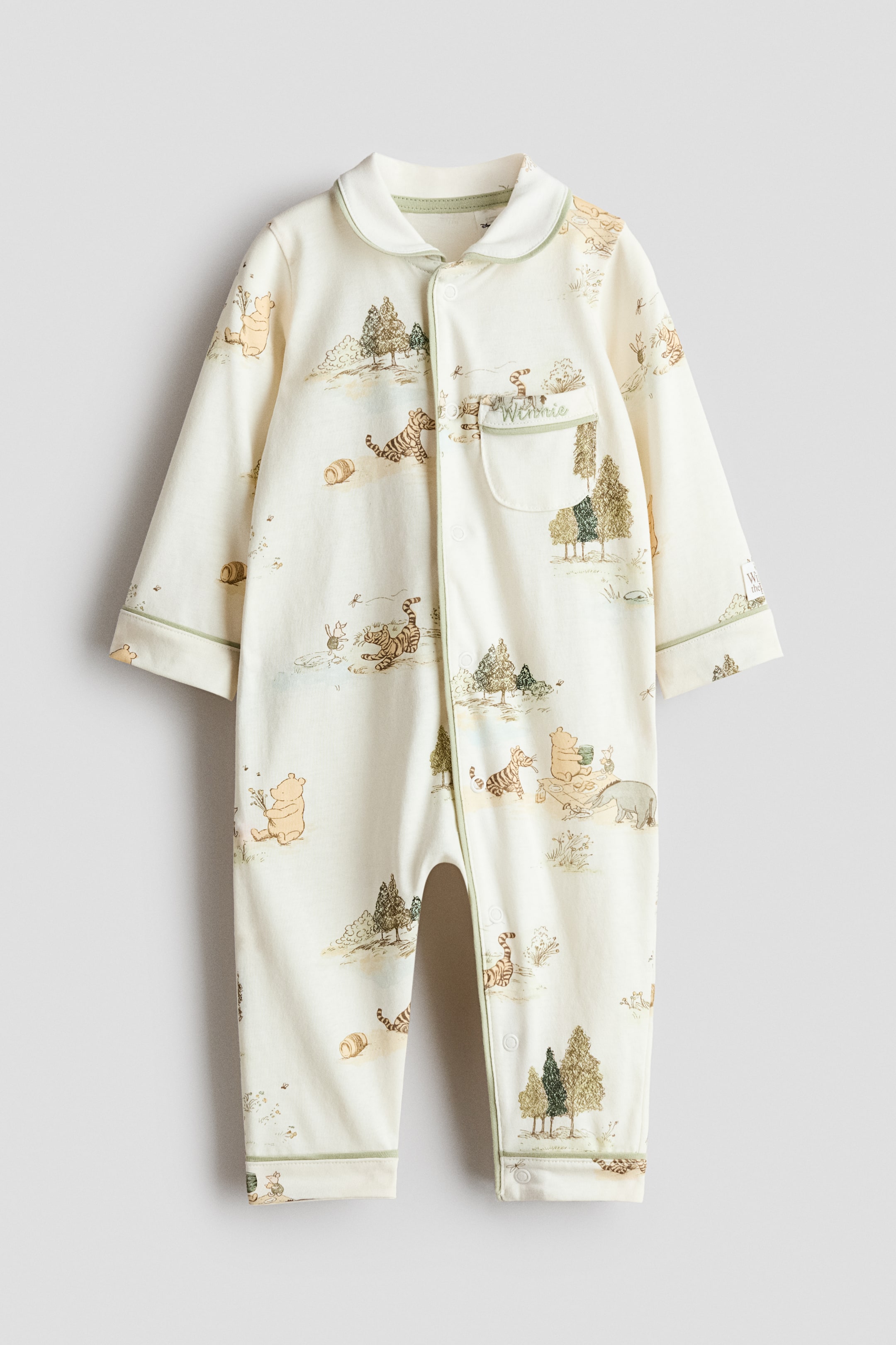 View larger image: A cream-coloured baby sleepsuit is hanging flat, showcasing a Winnie the Pooh and friends nature scene print. It is crafted with a rounded Peter Pan collar and green piping detailing the collar, front placket, cuffs, and ankle hems. A full snap button closure runs down the front and inner legs, and a small patch pocket with "Winnie" embroidered is visible on the left chest.