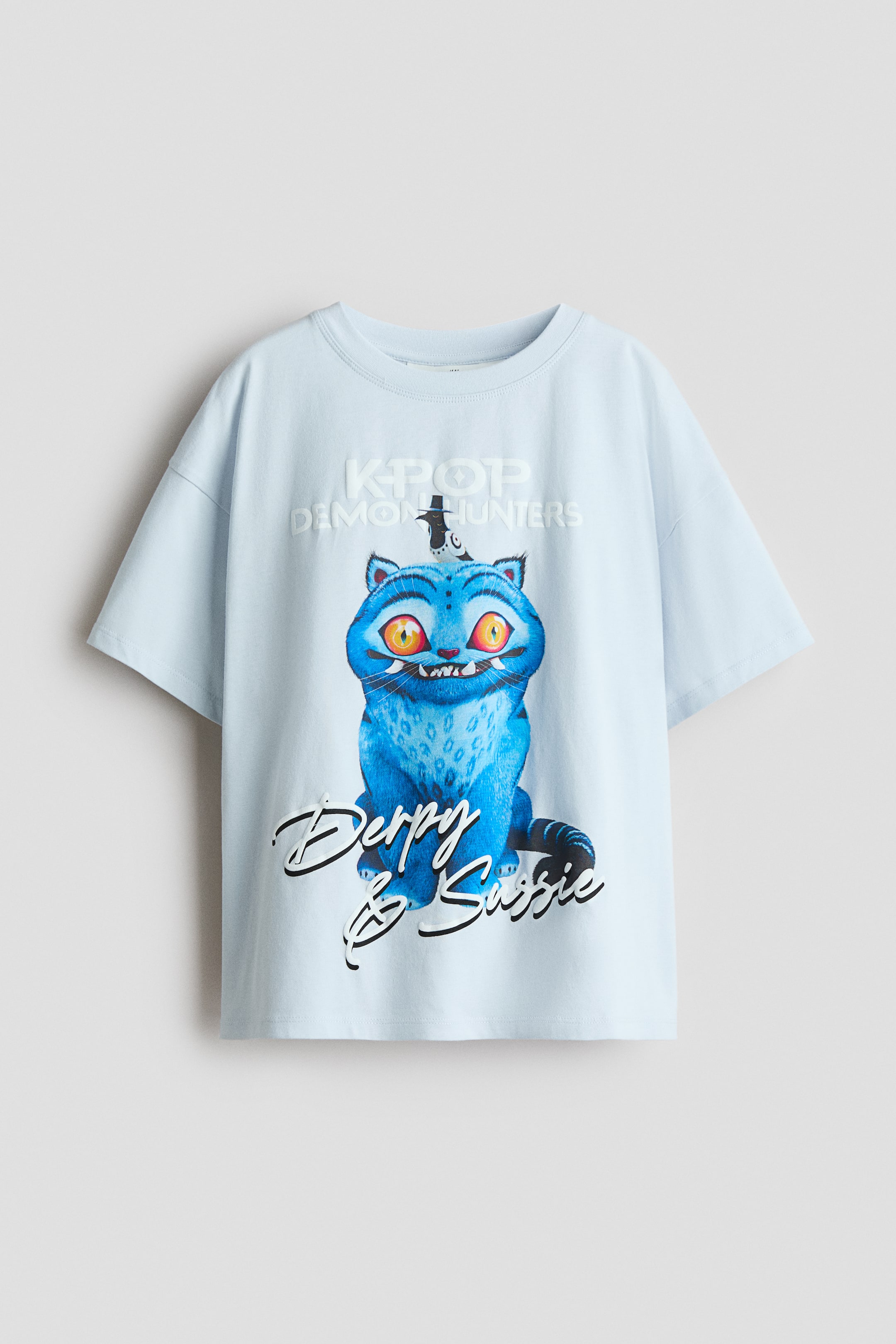 Se større billede: En lyseblå t-shirt lavet af glat jersey, har en rund, ribkantet halsudskæring og sænkede skuldre. Foran er der et stort printmotiv af en blå, tegneserieagtig kat med gule øjne og skarpe tænder, iført en lille sort hat, placeret over teksten "Derpy & Susie" i sort skrift, og "KPOP DEMON HUNTERS" i præget hvid tekst over katten.