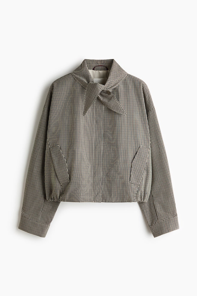 A cropped jacket showcases a black and off-white gingham pattern, with a large pointed collar tied into a bow at the front. It features two angled patch pockets, long sleeves with buttoned cuffs, and a gathered bottom hem. - 1