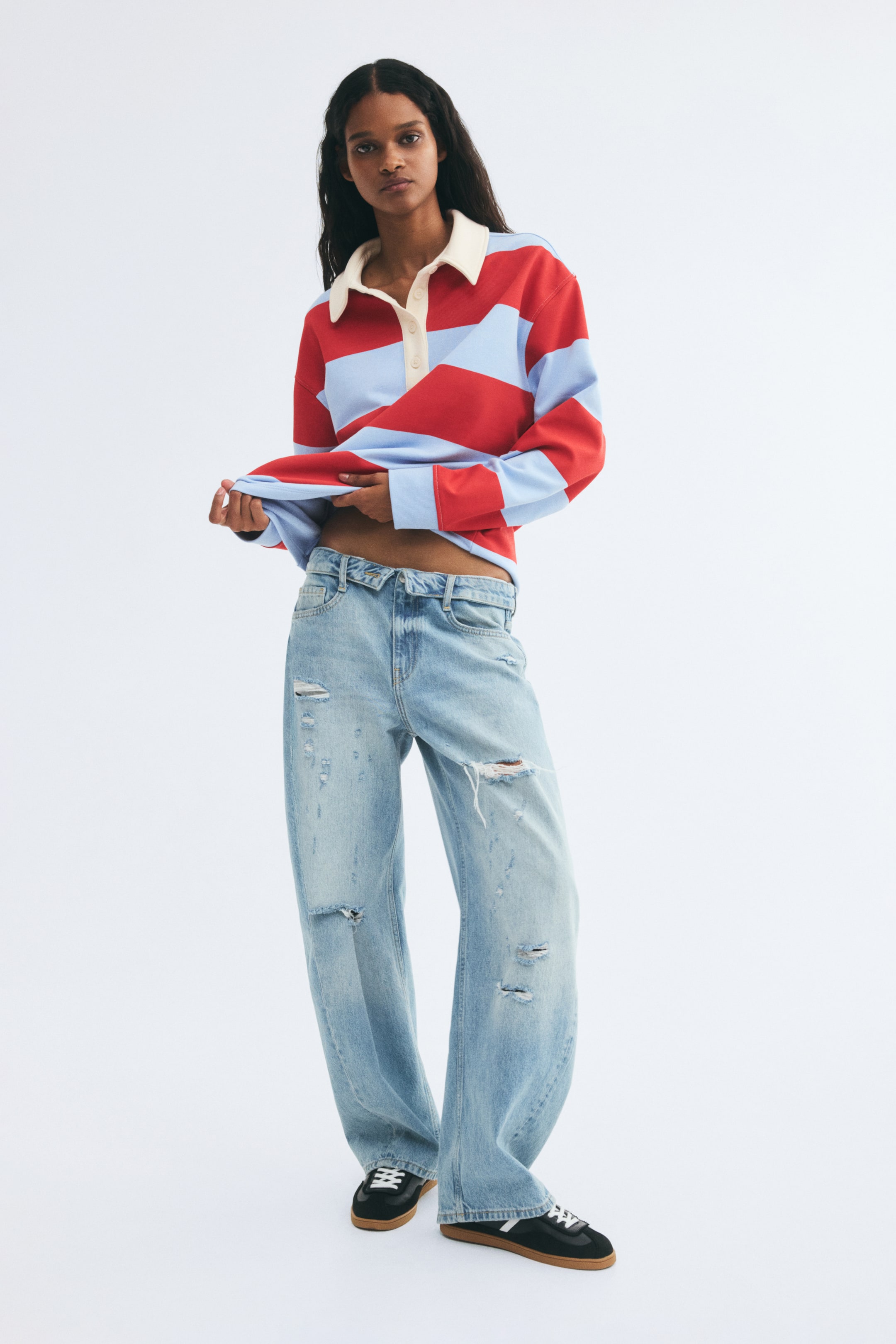View larger image: A loose-fit rugby shirt featuring bold red and light blue horizontal stripes, a contrasting cream collar, and a button placket is being worn by the model. The model is holding up the straight hem of the shirt with both hands, styling it with light-wash ripped jeans and black sneakers.