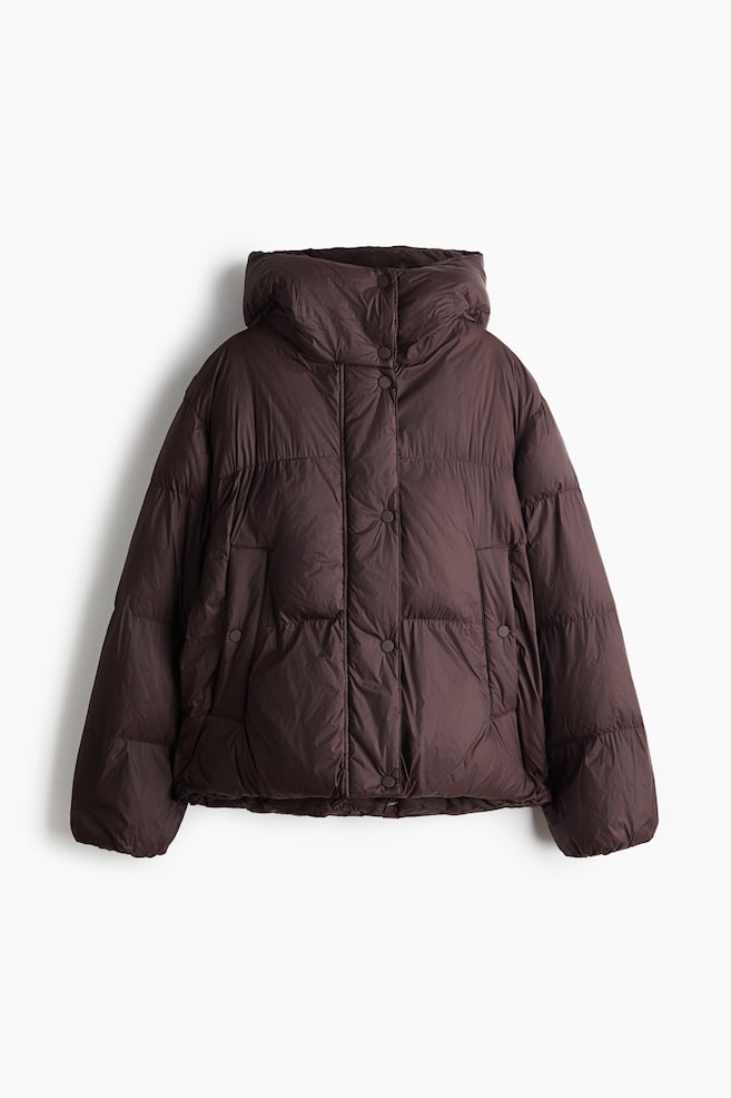 A dark brown puffer jacket is facing front, featuring horizontal quilting, a high padded collar with an integrated hood, and a central snap-button closure. The jacket is displaying long, voluminous sleeves and side welt pockets with snap details. - 1