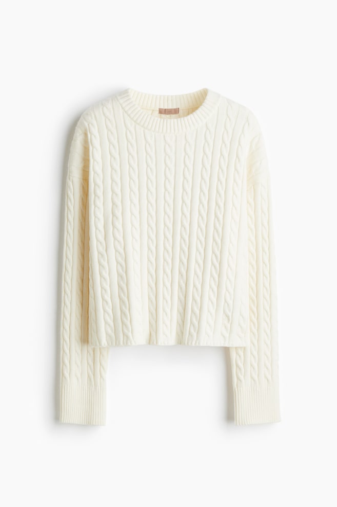 An off-white cable-knit sweater is being presented from the front, featuring a crew neck and long sleeves with ribbed cuffs. Its body and sleeves are showcasing a prominent cable knit pattern, while the bottom hem is also ribbed. - 1