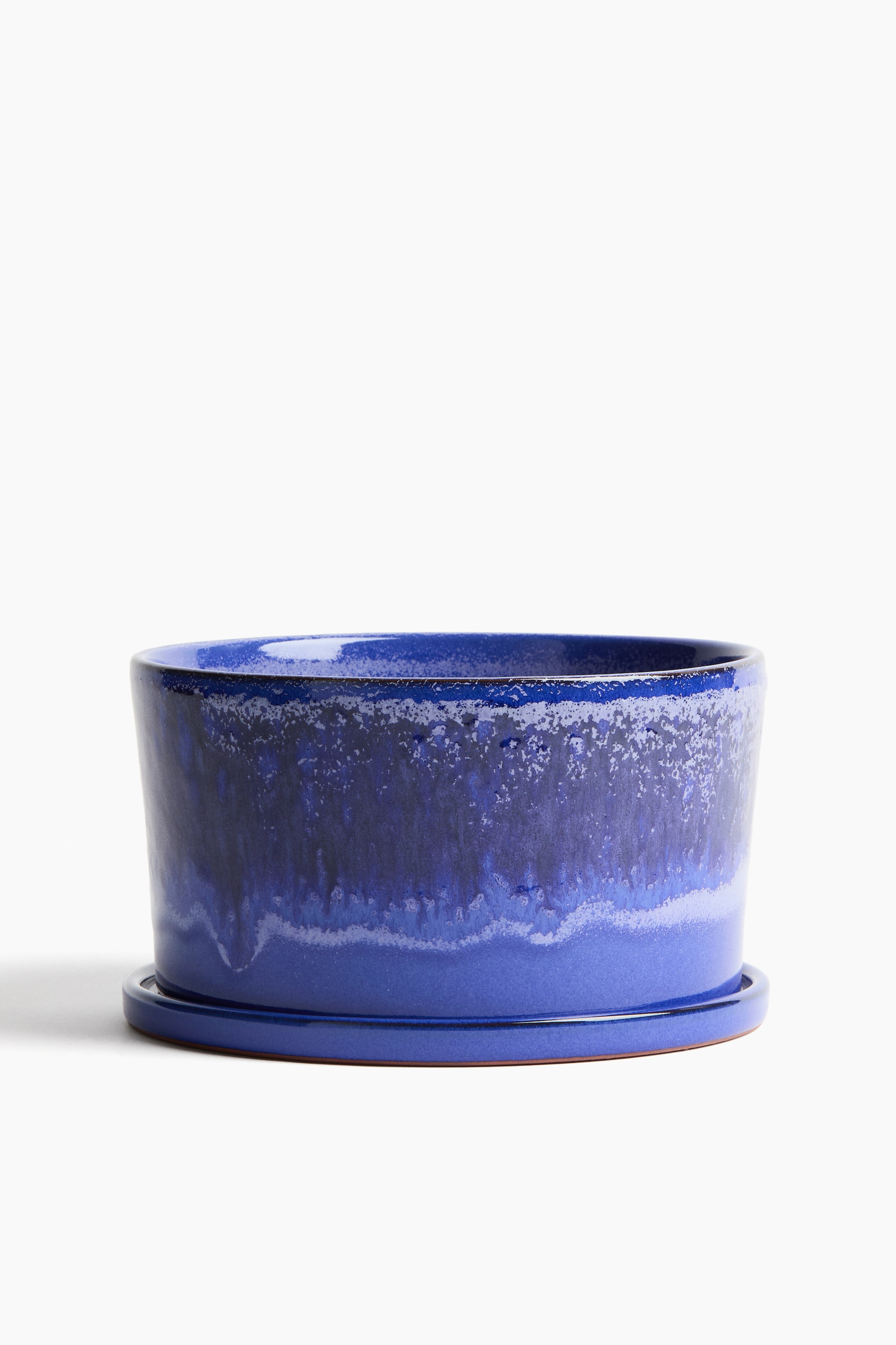 Visualizza immagine più grande: Un vaso per piante smaltato blu, caratterizzato da un design basso e largo con una smaltatura blu variegata che mostra toni più chiari e più scuri, è posizionato davanti su un sottovaso blu coordinato con un bordo non smaltato di colore marrone-rossiccio visibile.
