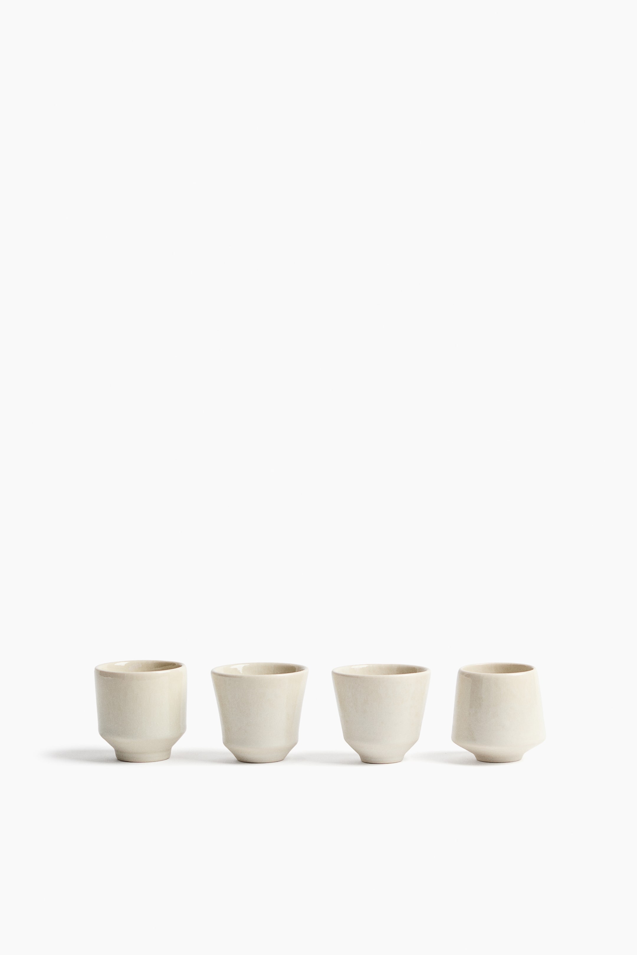 View larger image: Light beige ceramic cups showcase distinct, irregular forms. Each of the four cups features a unique shape, including a straight-sided design, a gently flared body, and an asymmetrical, angled form, all tapering toward the base.