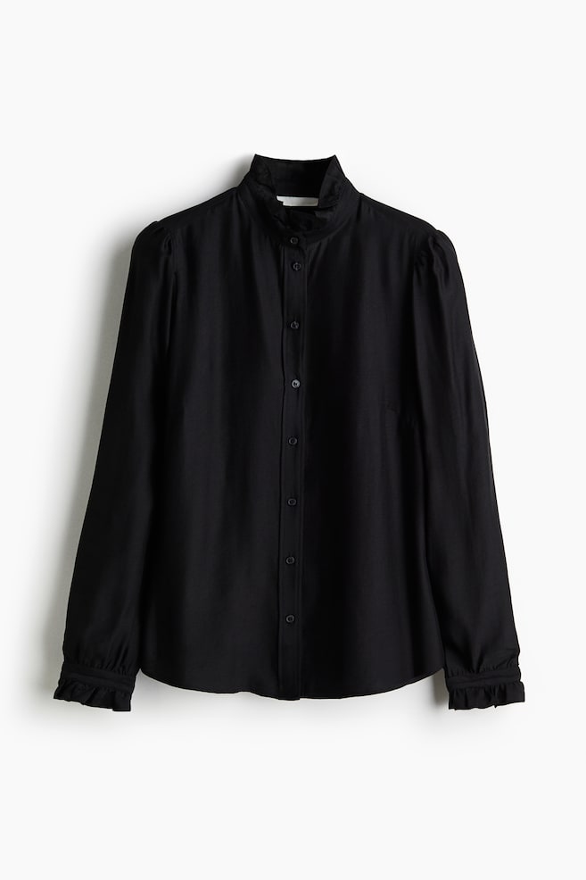Une blouse noire est présentée de FACE, avec un col à volants et des boutons sur le devant. Elle a de longues manches bouffantes avec des poignets à volants et un bouton, et un ourlet arrondi. - 1