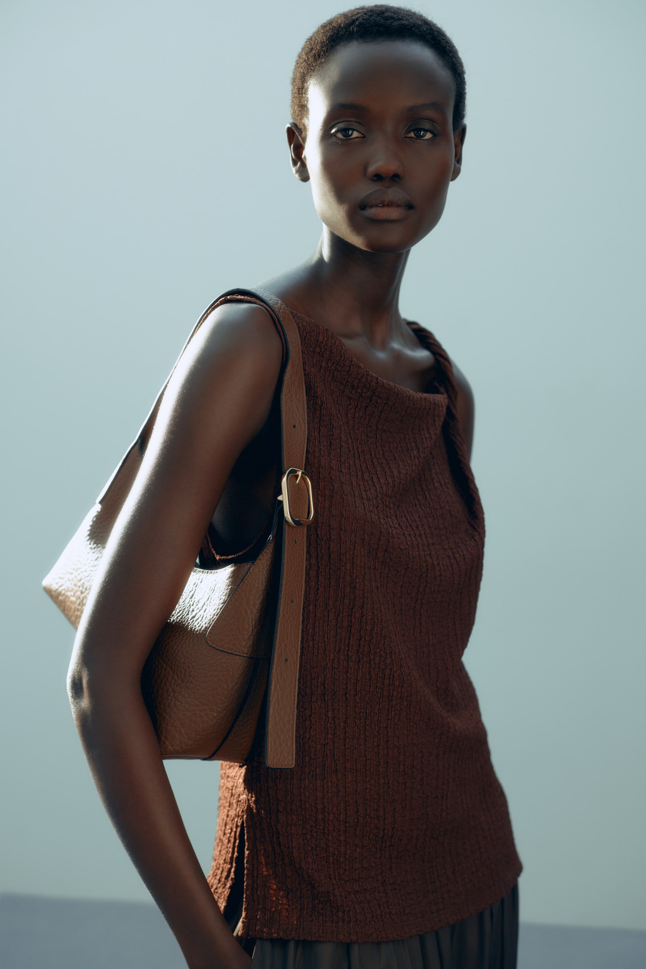 Xem ảnh lớn hơn: Một chiếc áo không tay, ôm sát màu nâu bằng chất liệu jersey có kết cấu đang được người mẫu mặc, nổi bật với đường viền cổ bất đối xứng và dây đeo vai xoắn tạo hiệu ứng xếp nếp nhẹ nhàng. Một chiếc túi xách vai màu nâu đang được đeo qua chiếc áo.