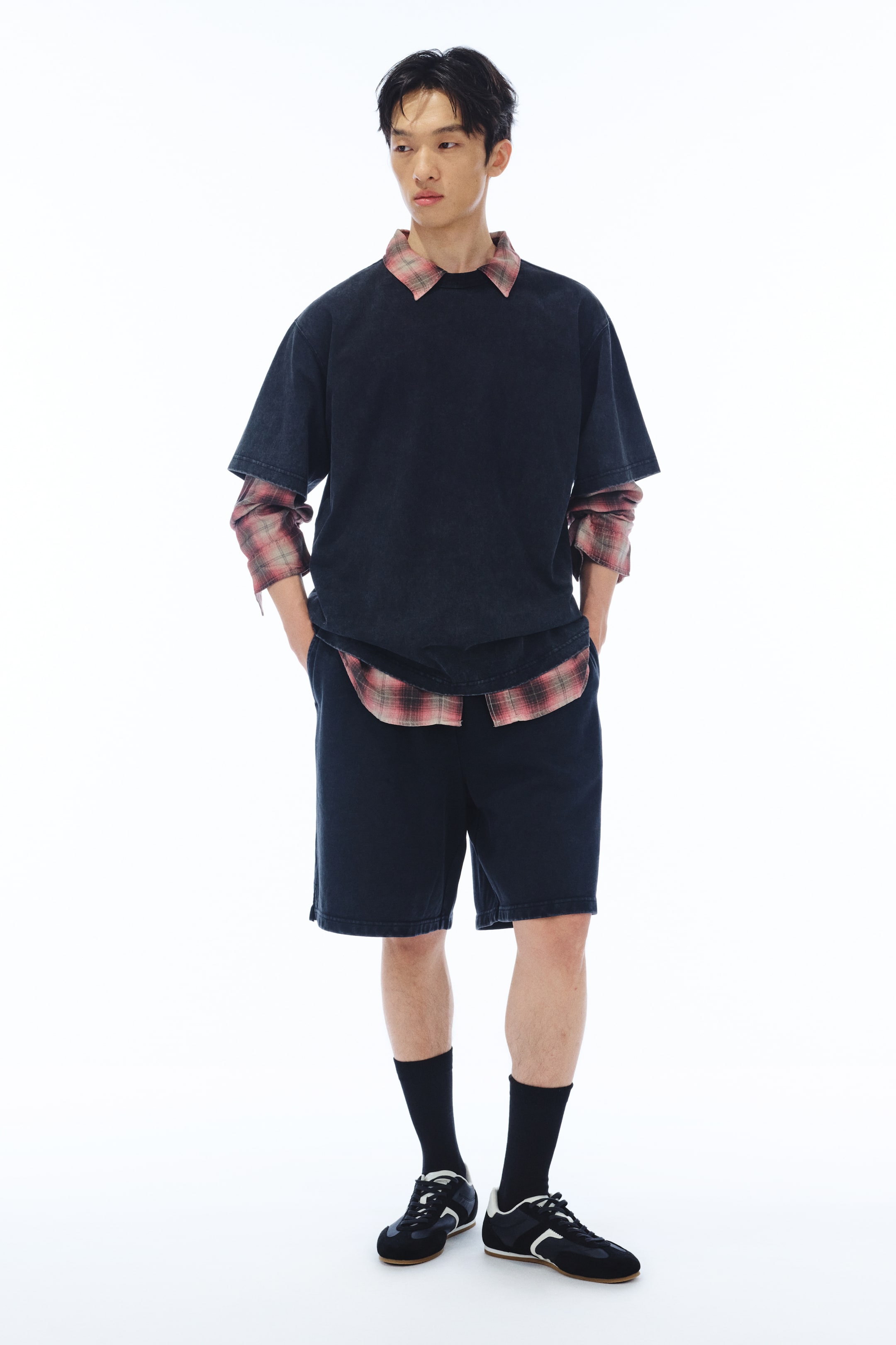 View larger image: The model is wearing an oversized black t-shirt layered over a red and black plaid long-sleeve shirt, with the collar, sleeves, and curved hem visible. Paired with black knee-length shorts, the model casually places both hands in the shorts' pockets. Completing the look are black athletic shoes with white accents, worn with black socks.