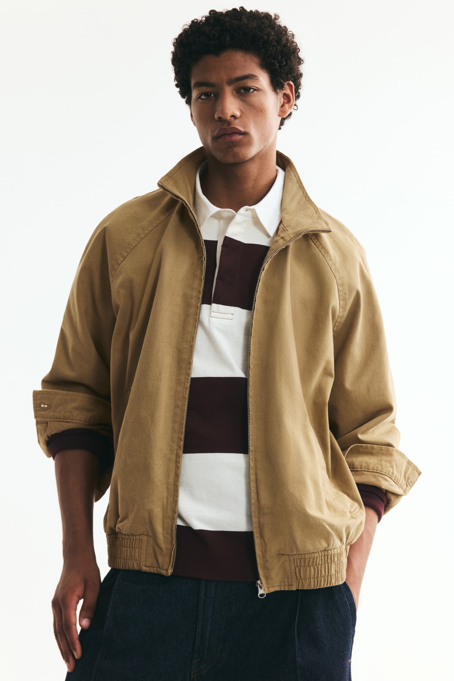 A front-facing tan jacket featuring a standing collar and a central zip closure. It's designed with two welt side pockets, long raglan sleeves with cuffs, and an elasticated hem. - 1