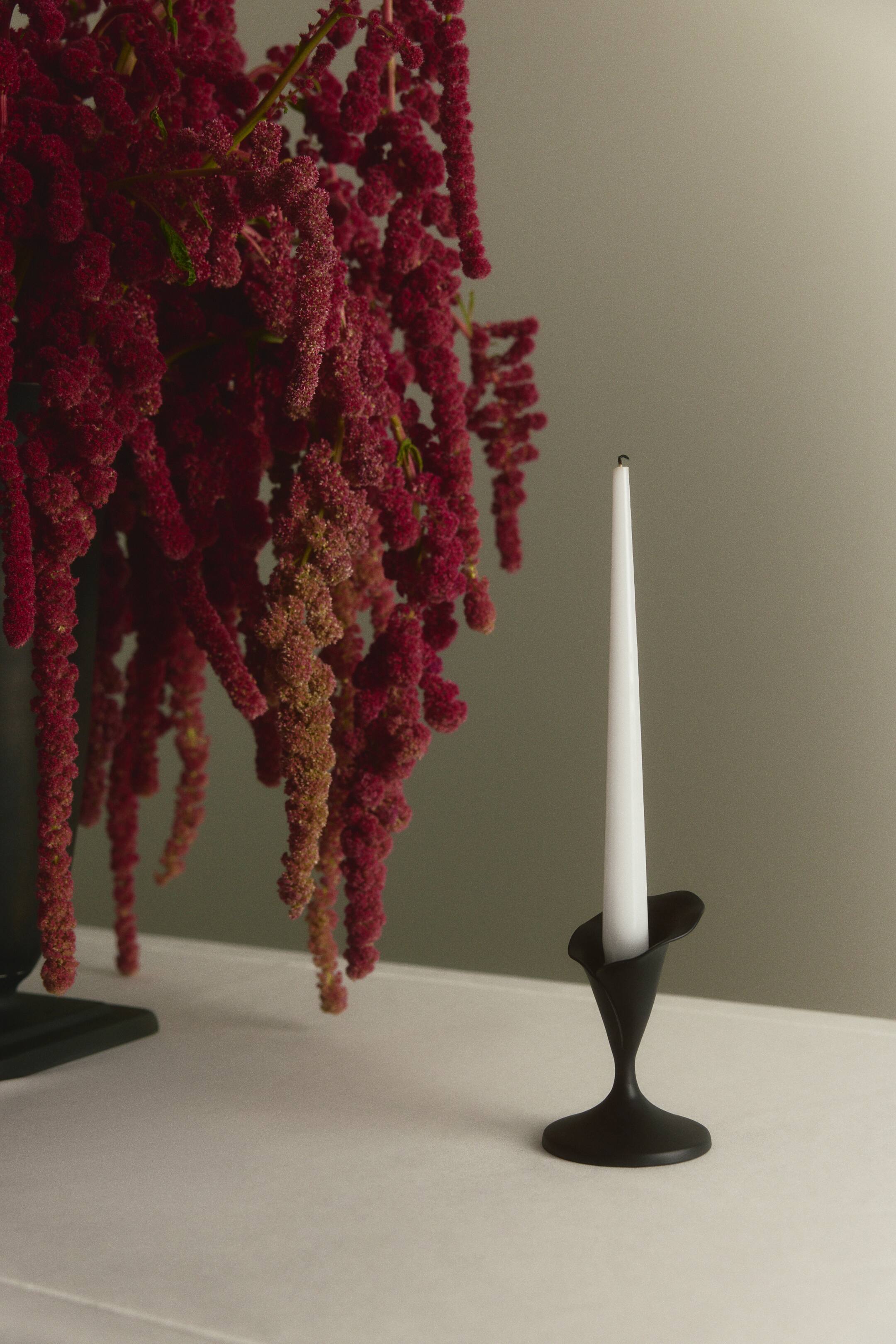 View larger image: A matte black metal candlestick featuring a flower-shaped socket, a slender stem, and a wide circular base holds a tall white taper candle. The candlestick stands on a flat surface, shown from a front-facing angle to highlight its sculptural silhouette.