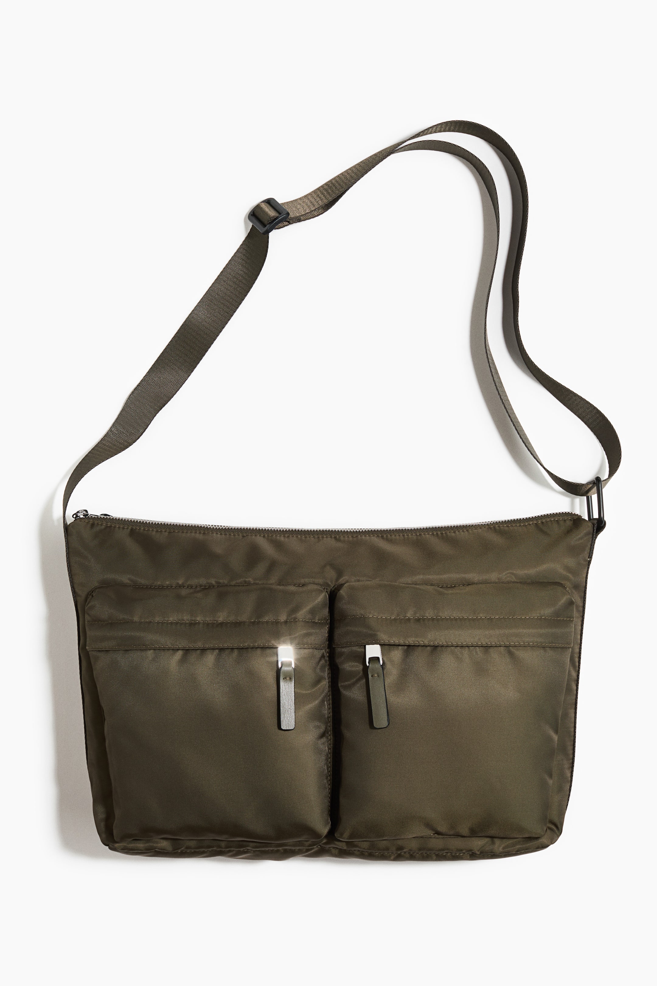 View larger image: An olive-green nylon crossbody bag faces front, featuring an adjustable strap and a main zip-fastened compartment. Two front patch pockets display a silver-toned zip pull and an olive fabric pull tab.