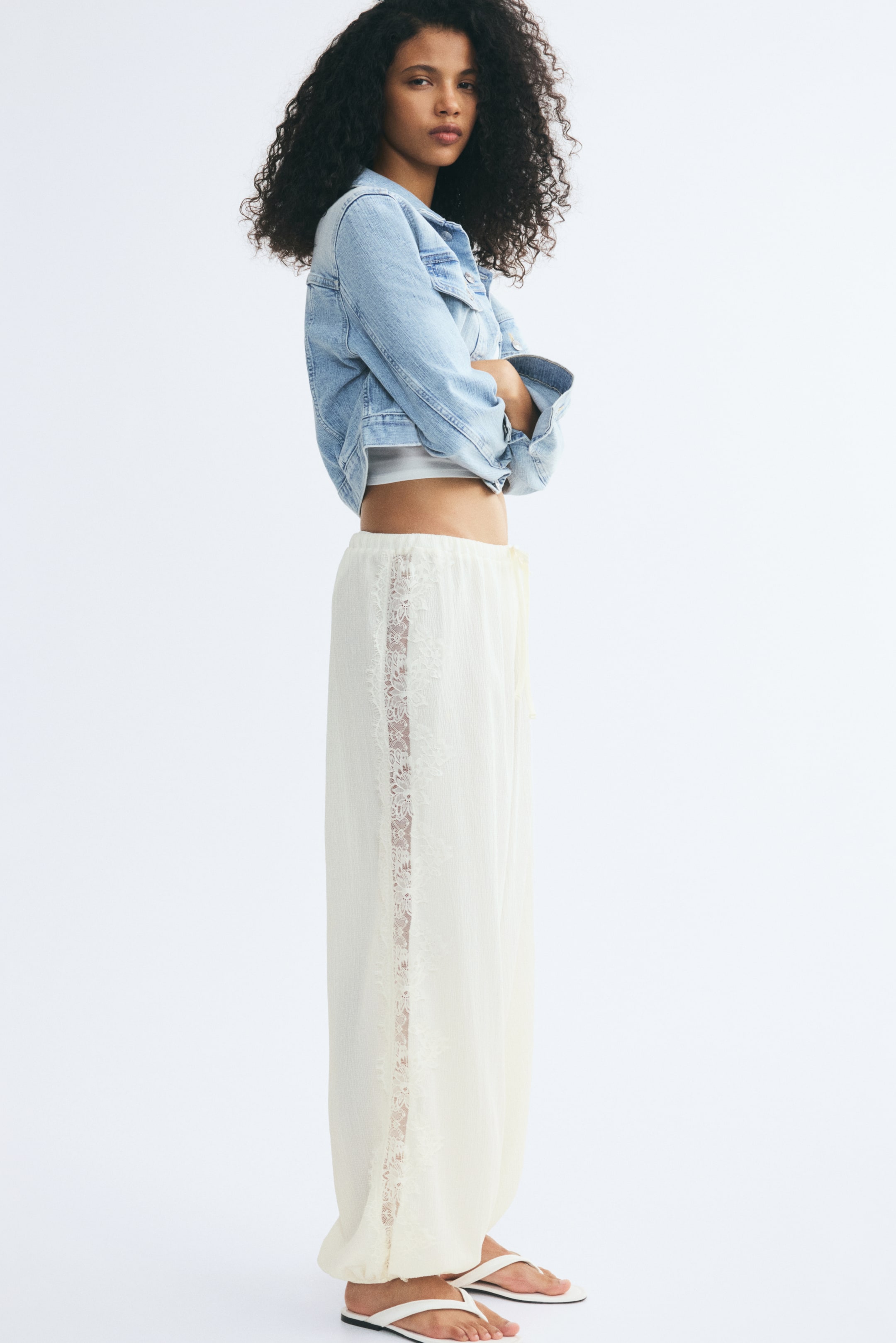 Ver imagen más grande: La modelo lleva unos pantalones de corte holgado en jersey arrugado de color crema, con perneras tipo balloon y un delicado ribete de encaje a lo largo del lateral, además de bajos elásticos. Los pantalones se combinan con una chaqueta vaquera cropped, un top corto blanco y sandalias planas.