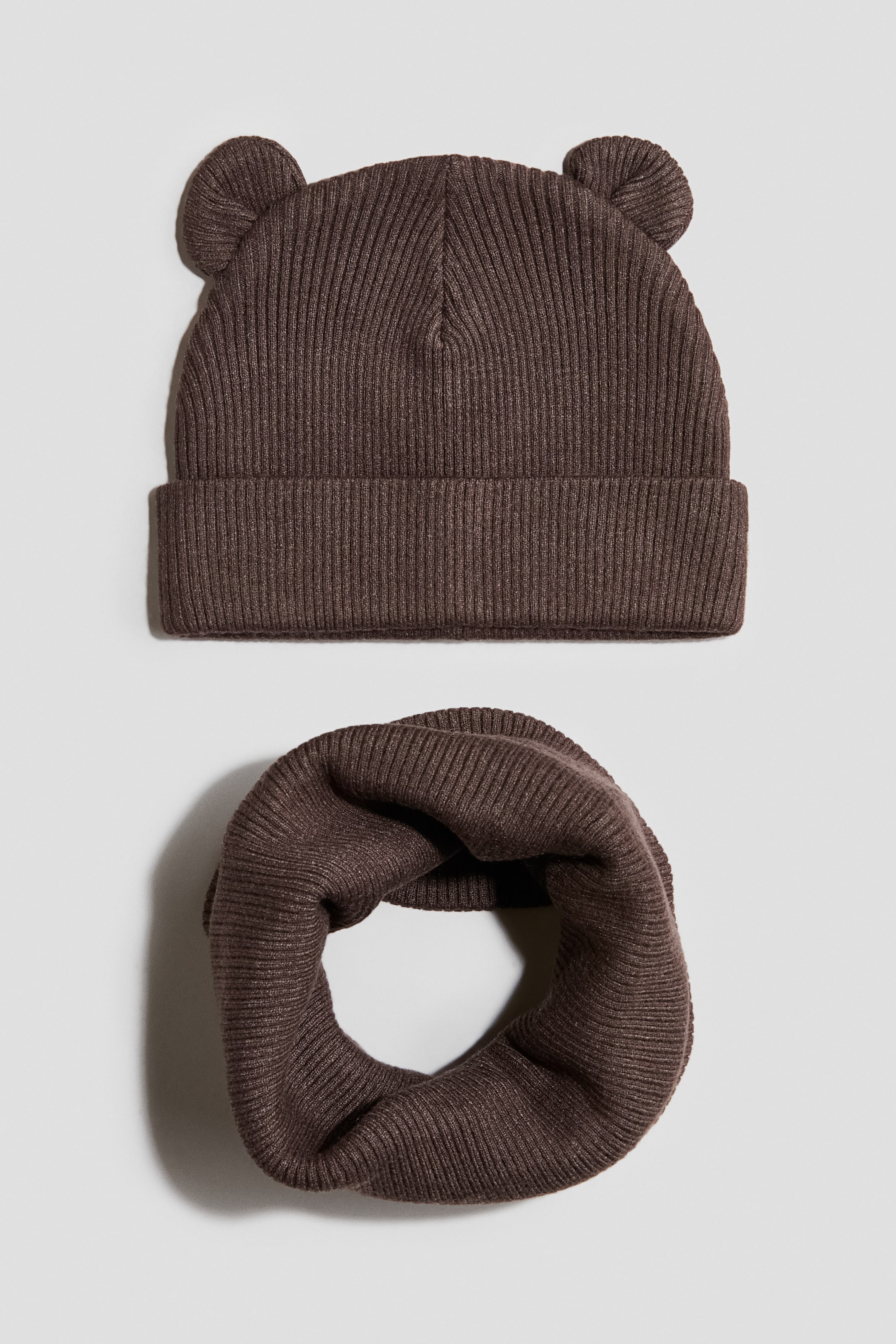 View larger image: A dark brown beanie and scarf set is displayed with a matching ribbed-knit texture, with the beanie facing forward and featuring a folded cuff plus two small round ears on top. Below the beanie, the matching infinity scarf rests in a loose circular loop.