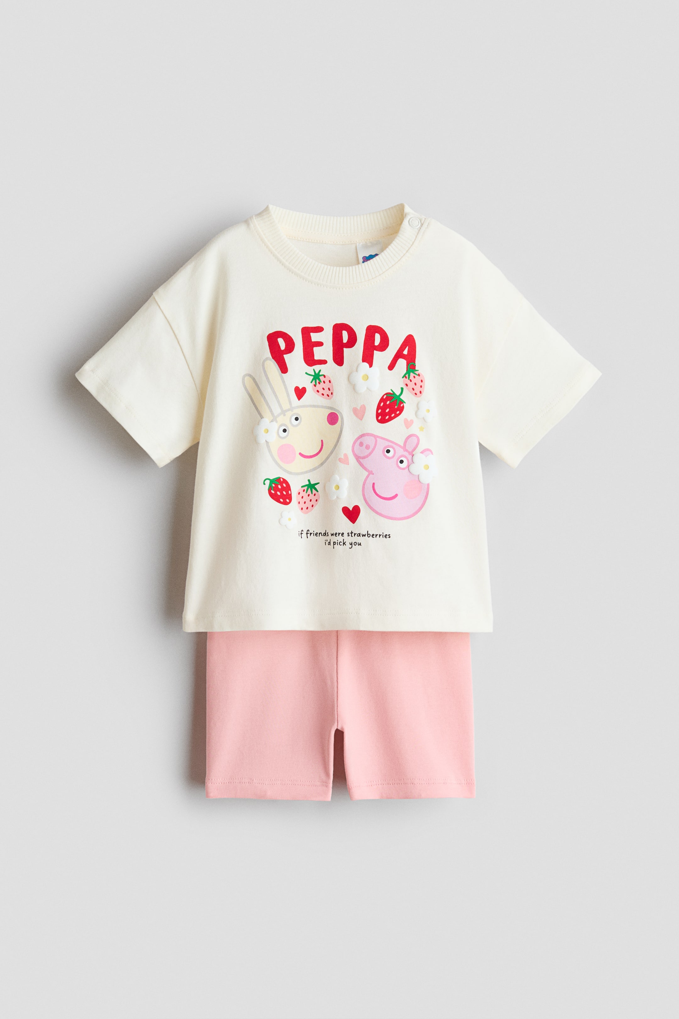 拡大画像を表示: 2ピースのプリントコットンセットが正面を向いています。クリーム色のTシャツとライトピンクのショーツが特徴です。Tシャツは、リブ付きのクルーネック、左肩に2つのスナップボタンがデザインされており、赤い「PEPPA」プリントと、ペッパピッグとウサギのキャラクターの顔、イチゴ、花、「友達がイチゴだったら、あなたを選ぶ」というテキストが描かれています。