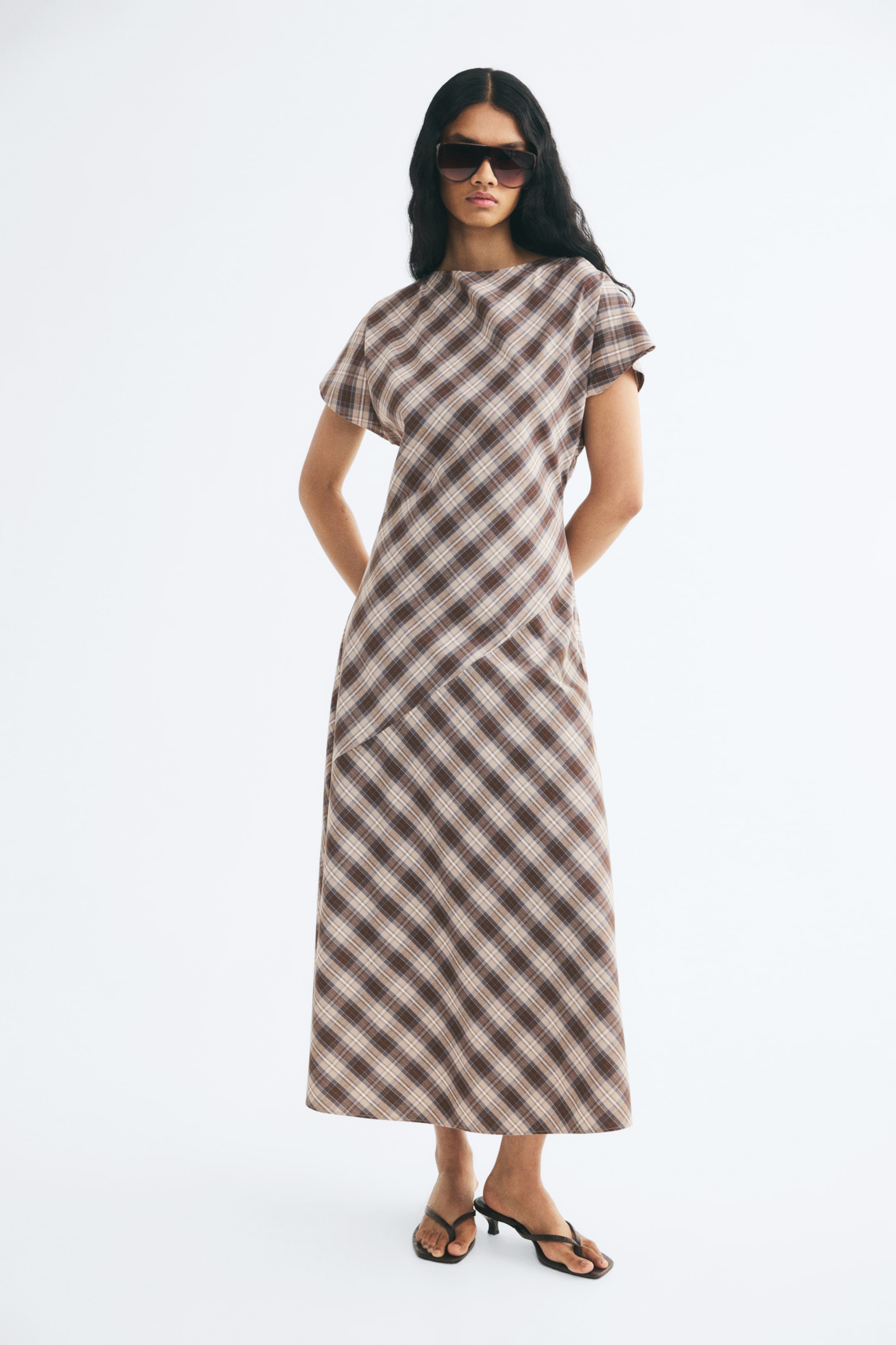 View larger image: A long, plaid woven dress in brown and beige is being worn by the model. The ankle-length dress features a boat neck, cap sleeves, and a prominent diagonal seam running across the hips.