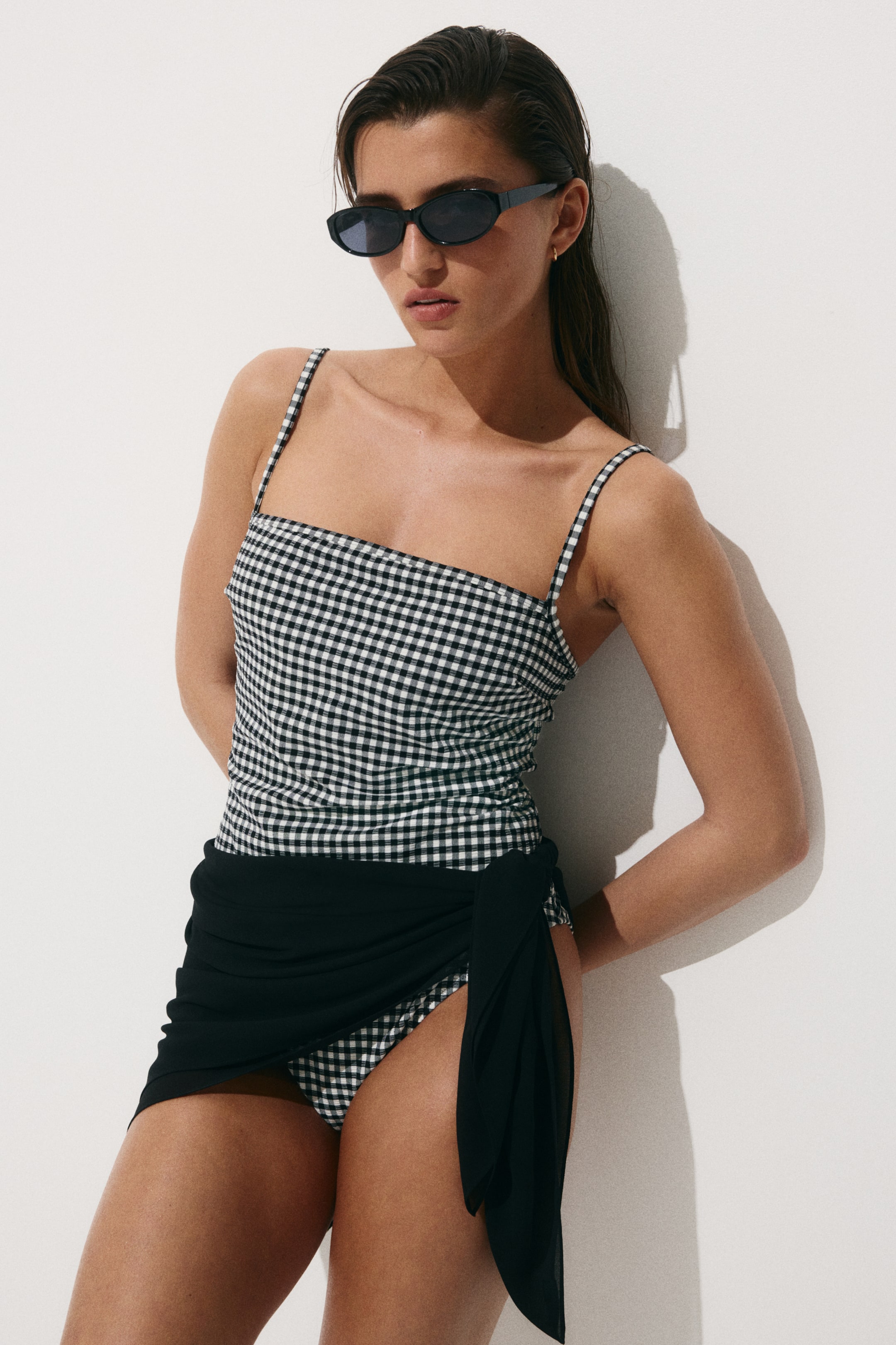 Vezi imaginea mai mare: Un costum de baie cu carouri gingham alb-negru este prezentat dintr-un unghi ușor frontal, având un top cu decolteu pătrat și bretele subțiri tip spaghetti, împreună cu slipuri asortate. Un sarong negru transparent este legat în jurul șoldului stâng, drapându-se peste slipul costumului de baie.