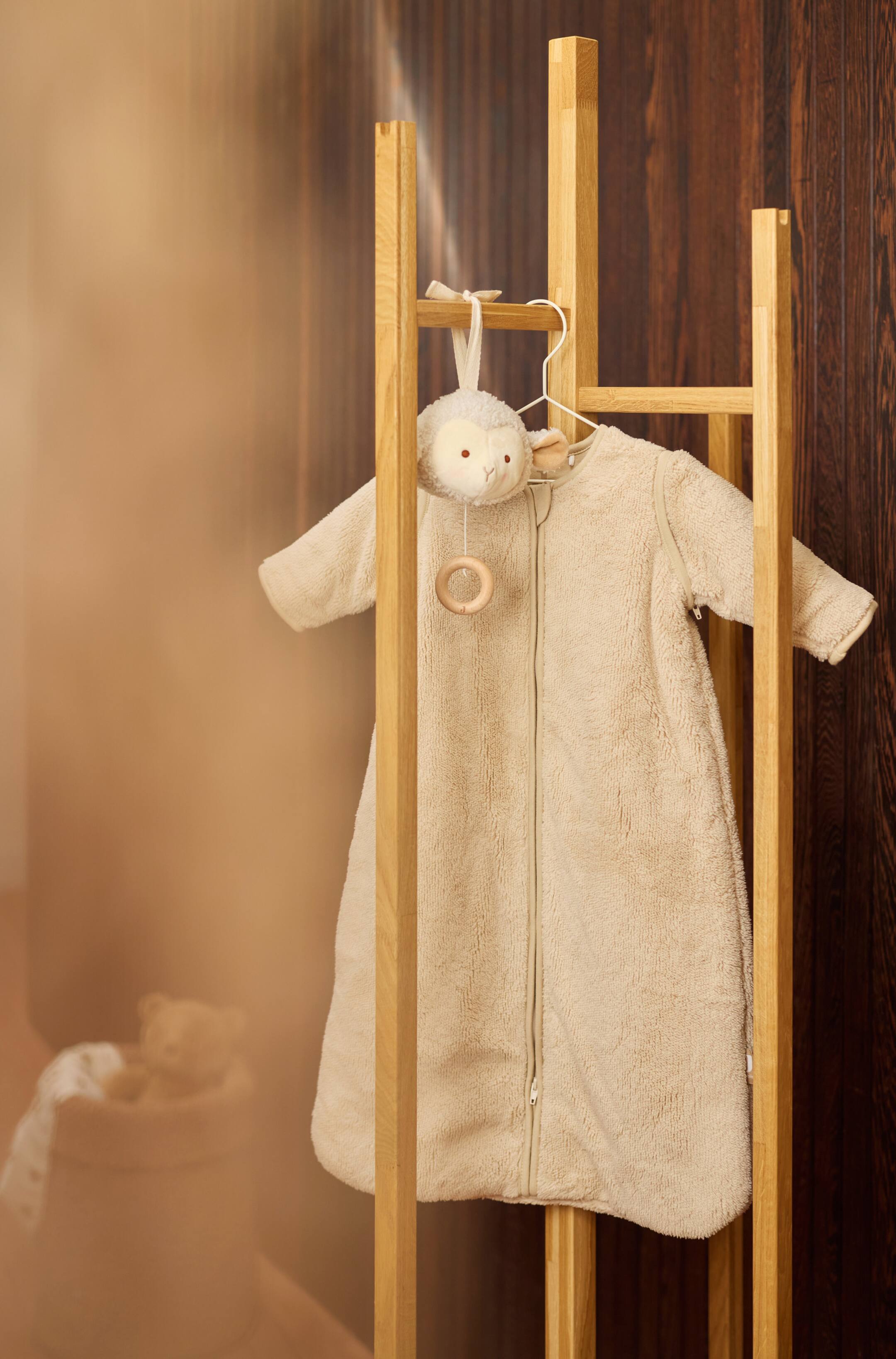 Größeres Bild anzeigen: Ein beiger, kuscheliger Babyschlafsack mit langen Ärmeln hängt nach vorne gerichtet an einem Holzständer und zeigt seine weiche Fleece-Struktur sowie einen durchgehenden Reißverschluss mittig vorne. Ein kleines, passendes Plüsch-Schaf mit einem Holz-Beißring hängt am oberen Ende des Reißverschlusses.