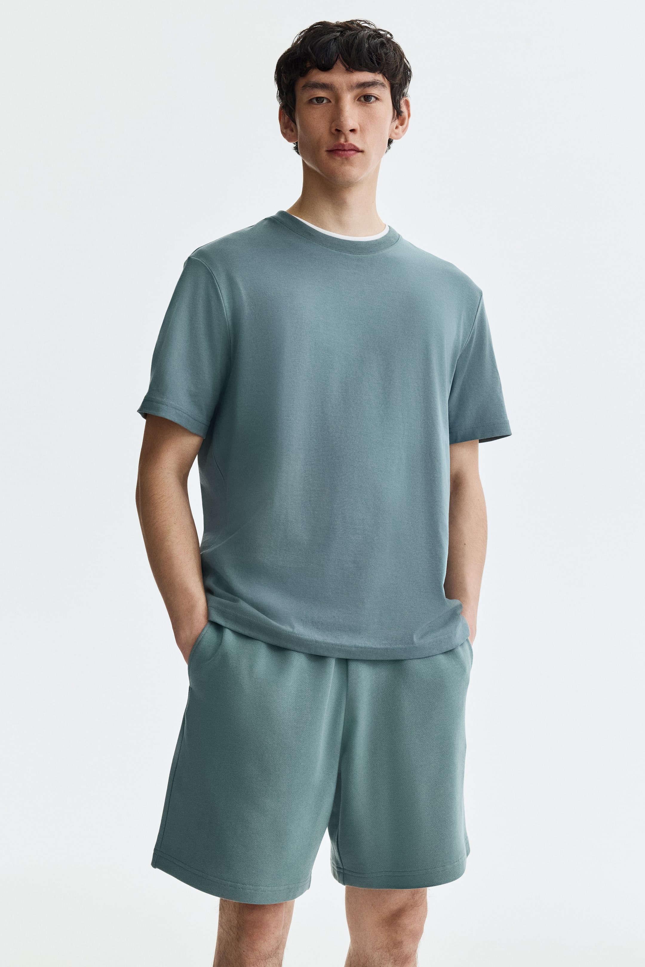View larger image: Teal sweatshirt shorts, featuring an elastic waistband and a regular fit, are worn by the model. The shorts include side pockets, with the model's hands placed inside.