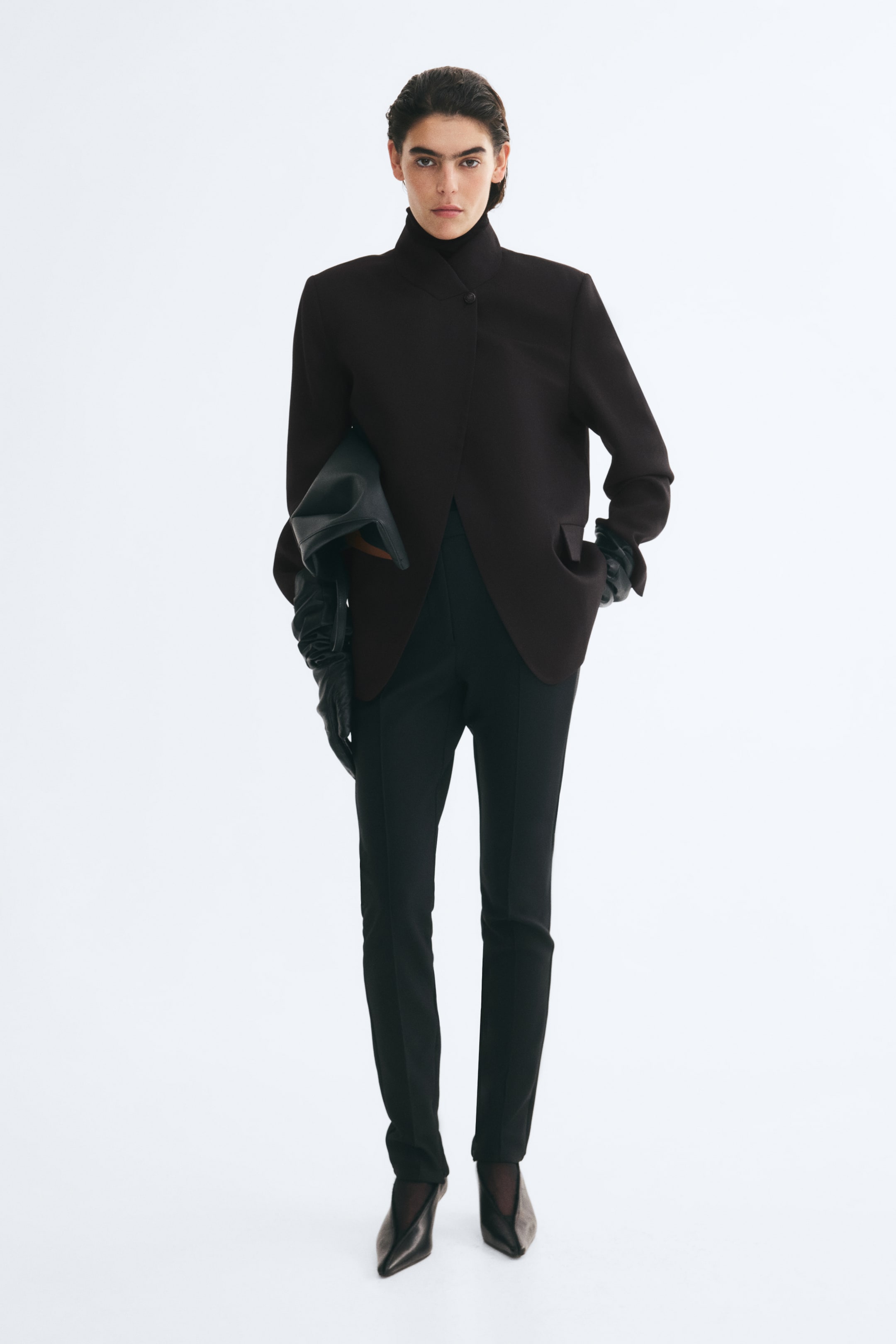 Se større billede: En oversized, mørkebrun vævet blazer bæres af modellen og har en høj ståkrave, markerede skuldre og en wrap-front-lukning med en enkelt synlig knap ved halsen. Blazeren er stylet med smalle sorte bukser, lange sorte læderhandsker og en sort clutch, der holdes under armen.