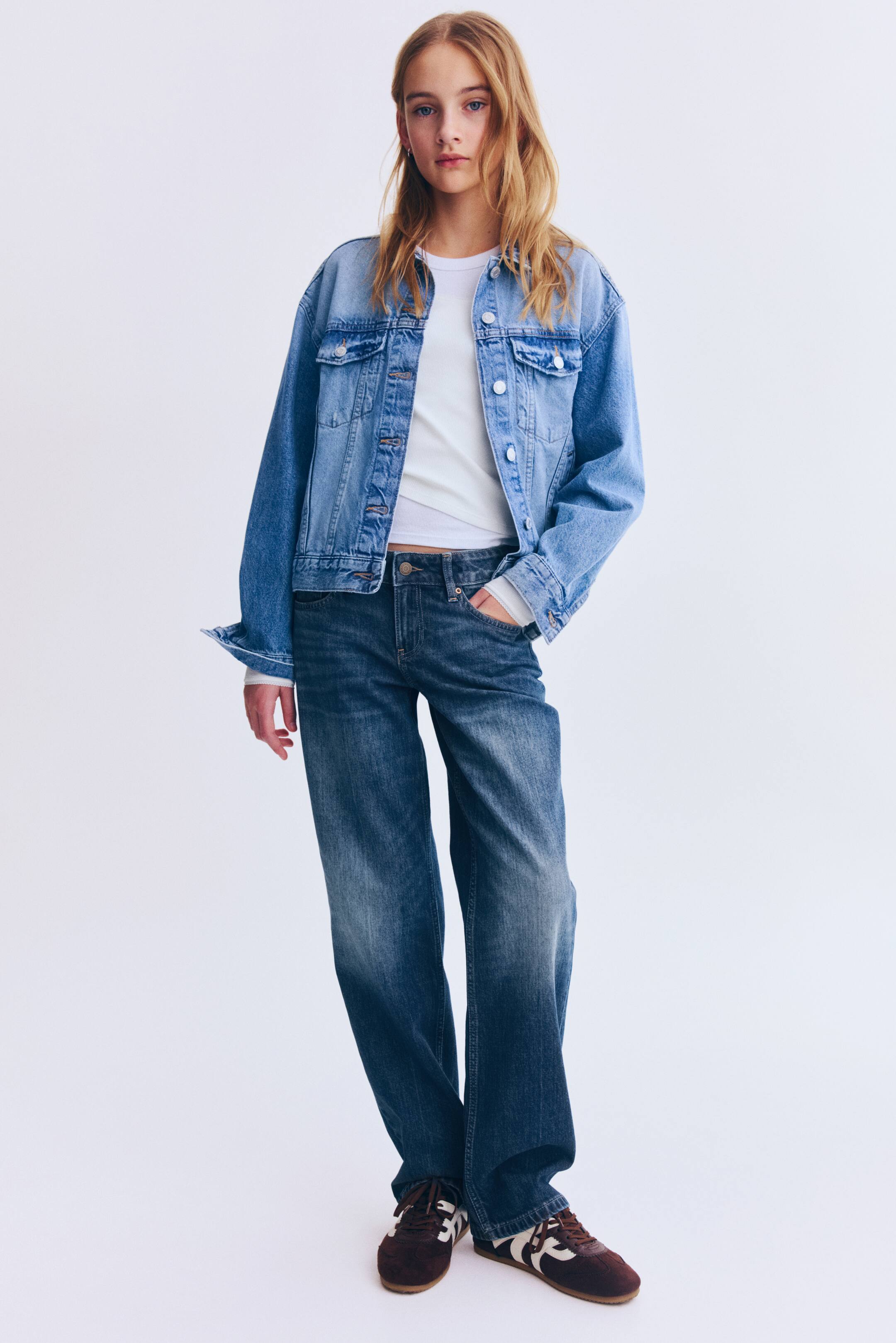 Преглед на по-голямо изображение: Широко кройано дънково яке в светъл деним с закопчаващи се джобове на гърдите е носено разкопчано върху бяла блуза с дълъг ръкав. Визията е завършена с дънки от тъмен деним с свободна кройка и кафяви велурени маратонки с бели ивици, всички заснети отпред.