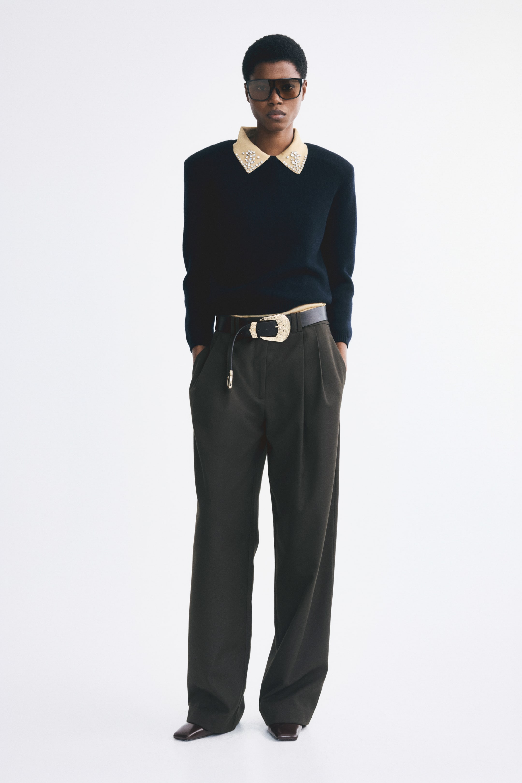 View larger image: Loose-fit, wide-leg pants in dark olive green woven fabric are worn by the model, who is standing with hands tucked into the side pockets. The pants feature front pleating and are styled with a black leather belt with a decorative gold buckle and a tucked-in black sweater.