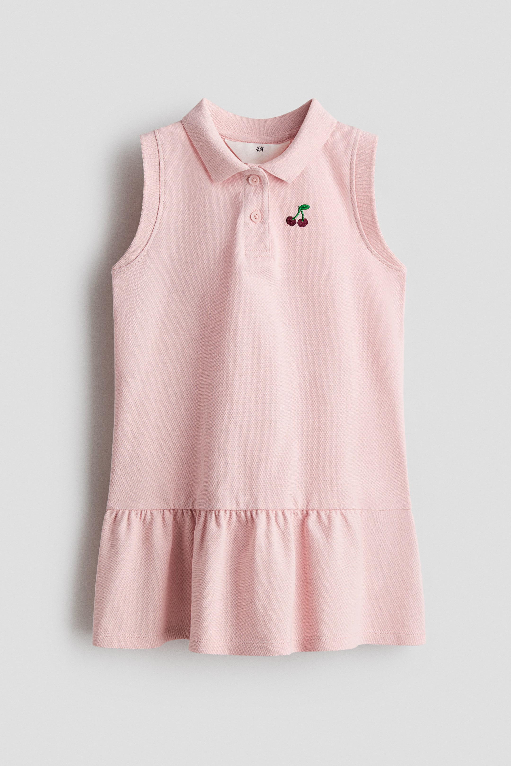 Ampliar la imagen: Un vestido tipo polo sin mangas rosa presenta la parte frontal, con un cuello polo, una tapeta de dos botones y un bordado de cereza en el pecho izquierdo. Un dobladillo con volantes forma la parte inferior del vestido.