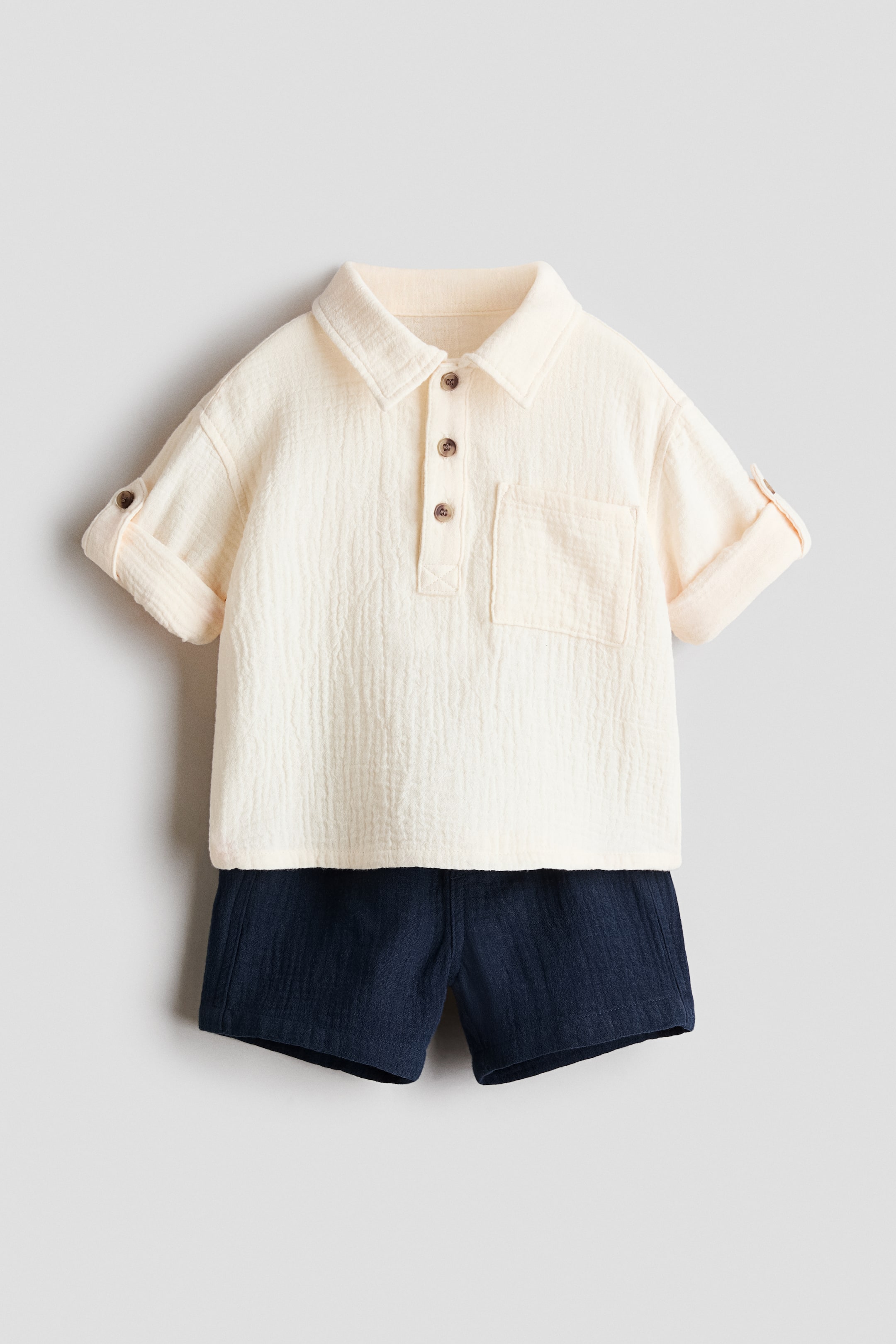 Voir une image plus grande: Un ensemble chemise en seersucker crème et short en seersucker bleu foncé est présenté de face. La chemise est dotée d'un col, d'une patte de boutonnage à trois boutons marron, d'une poche poitrine ouverte, d'épaules tombantes et de manches retroussables fixées par des pattes boutonnées. Le short présente une taille élastiquée.