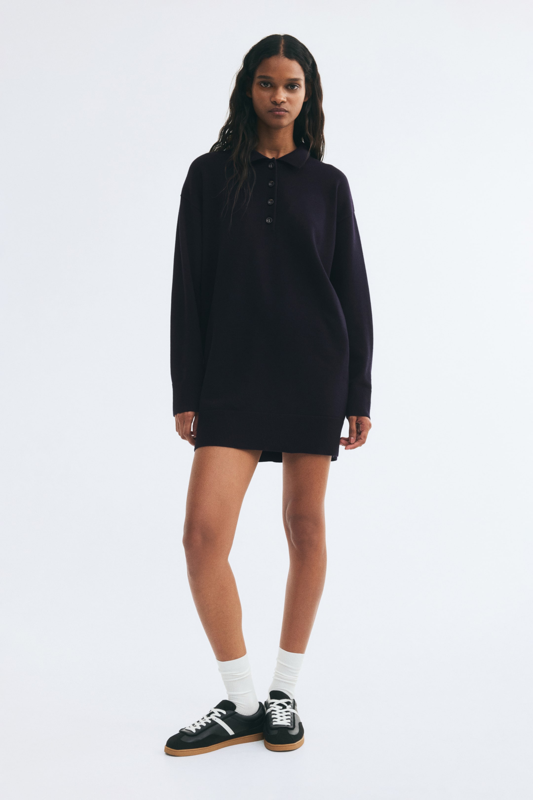Voir une image plus grande: Une courte robe en tricot violet foncé, dotée d’un col, d’une patte de boutonnage, de manches tombantes, ainsi que de poignets et d’un ourlet côtelés, est portée par le mannequin. Le mannequin accessoirise la robe avec des chaussettes blanches et des espadrilles noires.