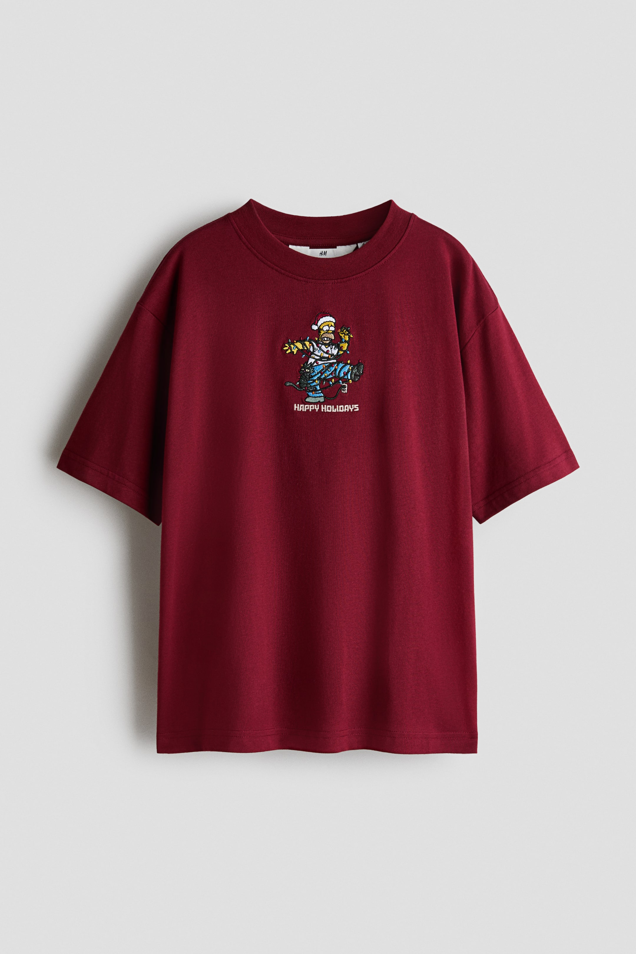 Voir une image plus grande: Un t-shirt en jersey de coton rouge foncé est présenté de face, avec un col rond à bord-côte, des épaules tombantes et un motif imprimé au centre de la poitrine. Le visuel représente Homer Simpson emmêlé dans des lumières de Noël colorées au-dessus du texte « JOYEUSES FÊTES ».