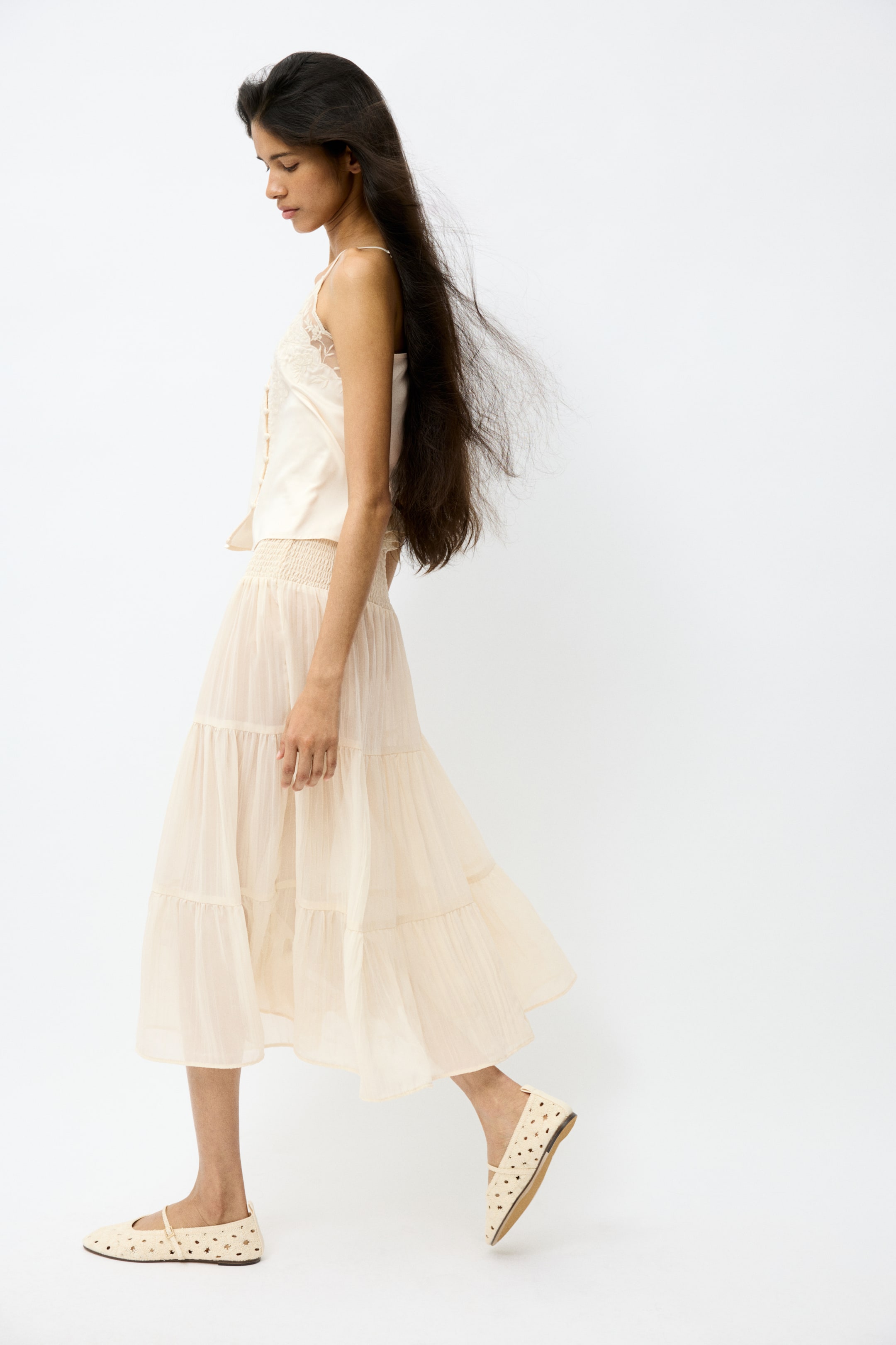 View larger image: A cream-coloured camisole top features lace trim along the v-neckline and a smocked hem, paired with a tiered maxi skirt that displays a wide smocked waistband and flowing layers. Light-coloured flat Mary Jane shoes, which feature a perforated design and an instep strap, complete the ensemble, all viewed from a side angle.