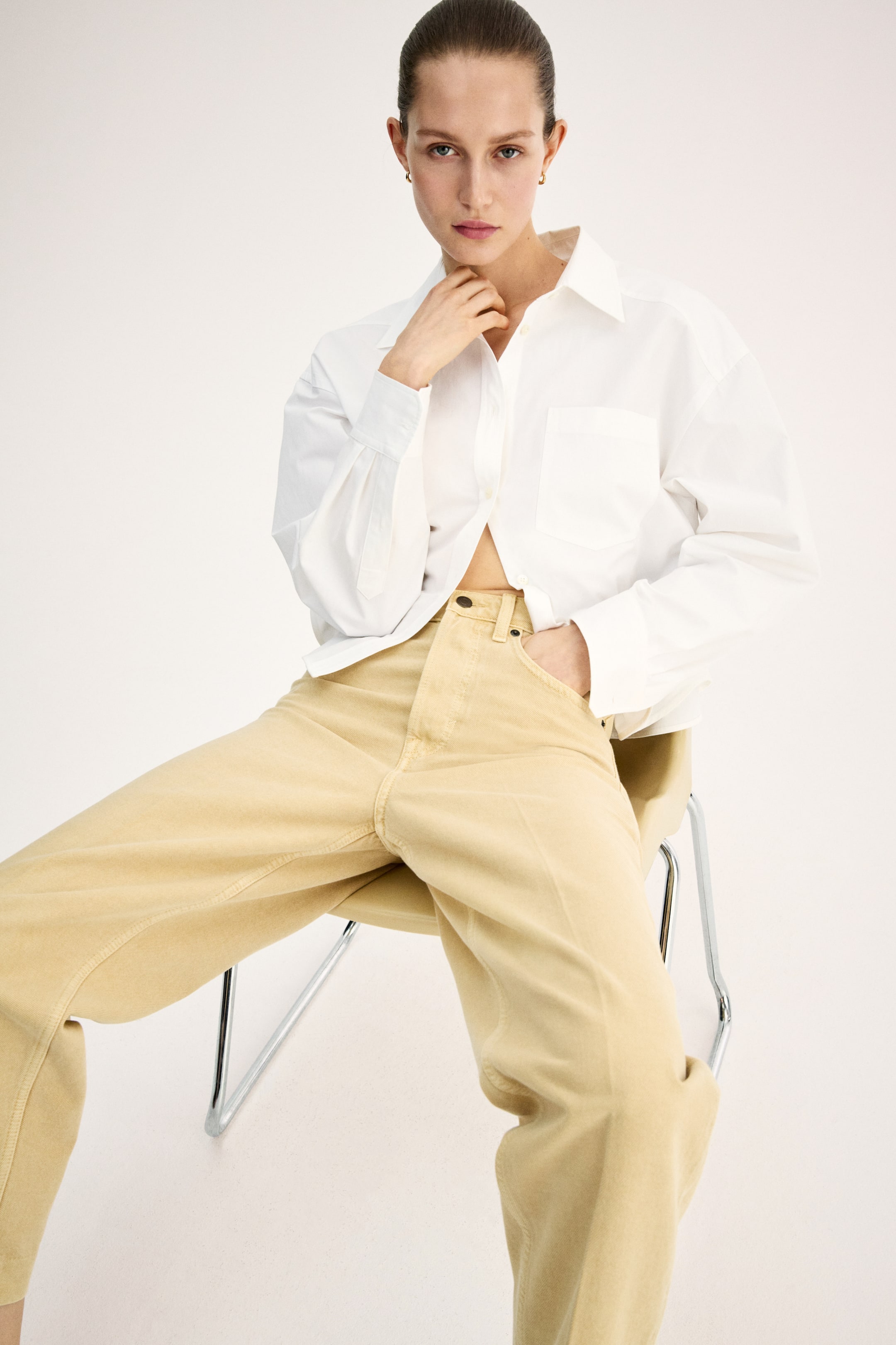 View larger image: Light beige 5-pocket denim jeans are worn by the model, featuring a high waist with a button fly, voluminous legs with visible front creases, tapering to an ankle length. The model is seated, with one hand resting inside a front pocket of the jeans.