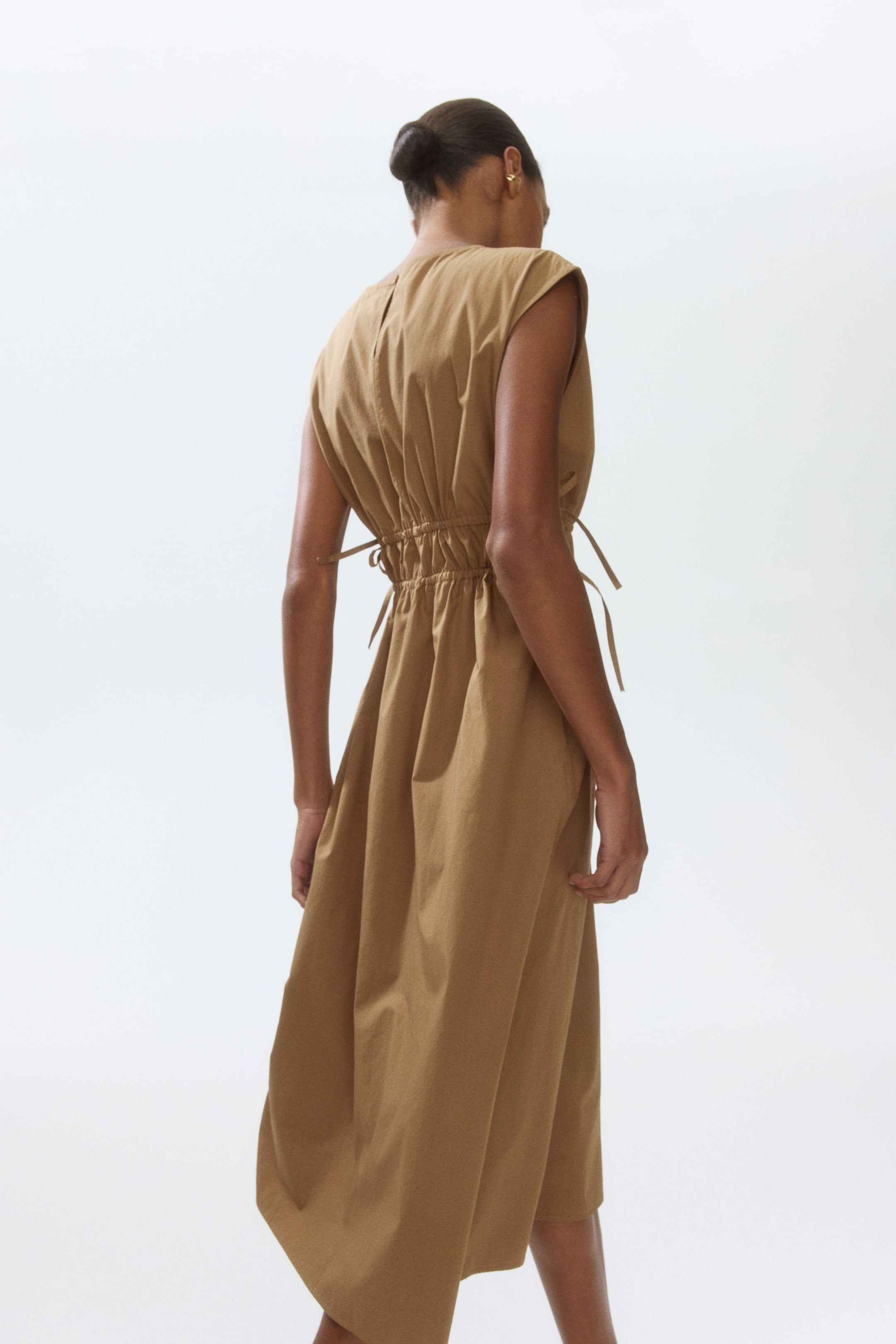 View larger image: The model is showcasing a long, sleeveless dress crafted from crisp tan cotton poplin. It features a round neckline with a keyhole opening and a fastening at the back. The fitted waist is elegantly cinched with two drawstrings, while the skirt gracefully flares out.