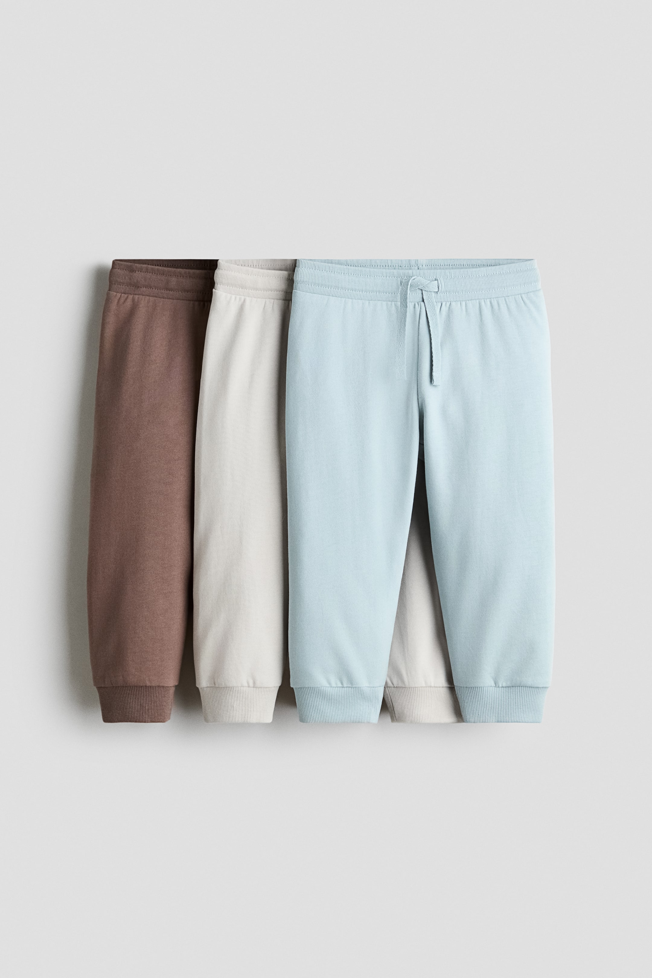 Grotere afbeelding bekijken: Drie paar katoenen sweattrackbroeken in bruin, lichtbeige en lichtblauw hangen in een overlappende rij, gezien van voren. Elk paar heeft een elastische geribde tailleband en geribde boorden aan de pijpen, waarbij het lichtblauwe paar aan de voorkant een bijpassend trekkoord in de taille heeft.