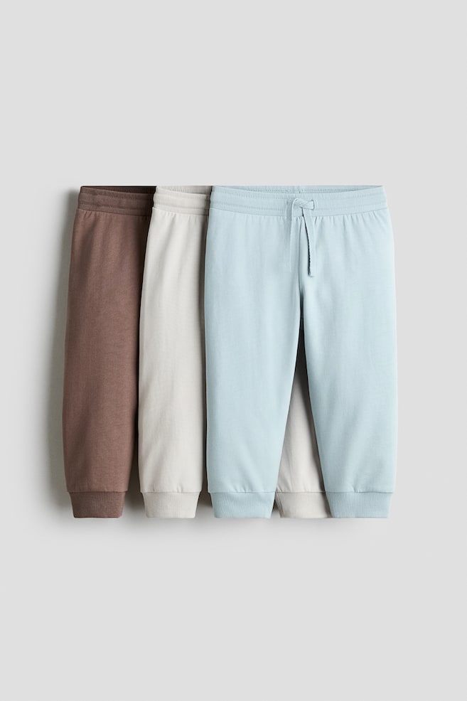 Three pairs of cotton sweat track pants in brown, light beige, and light blue are hanging in an overlapping row from a front view. Each pair features an elasticised ribbed waistband and ribbed cuffs at the hems, with the front light blue pair showing a matching drawstring at the waist. - 1