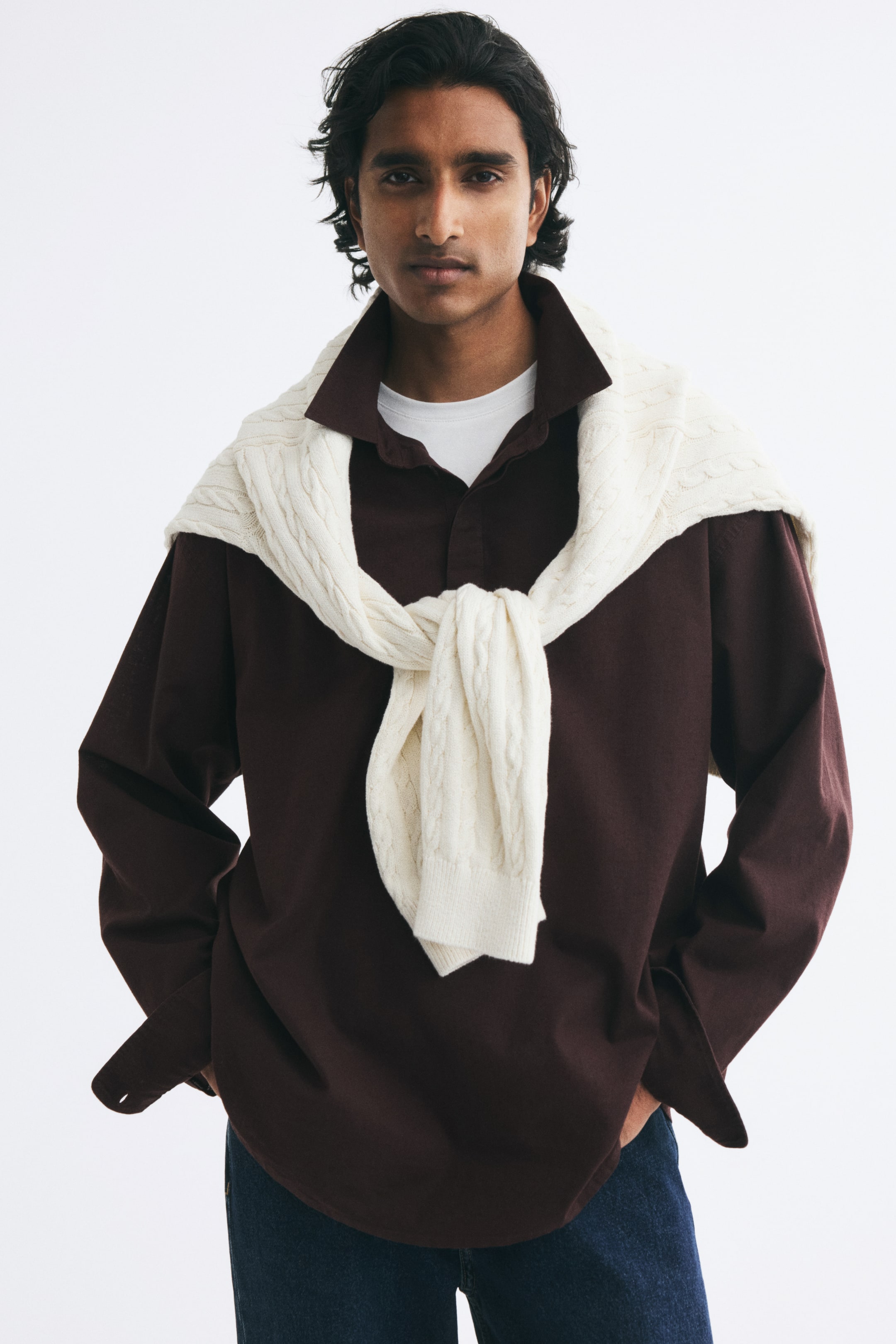 View larger image: The model is wearing a dark brown popover shirt layered over a white T-shirt and paired with blue jeans. A cream cable-knit sweater is draped over the shoulders and tied at the front, accentuating the shirt’s relaxed fit and collar.