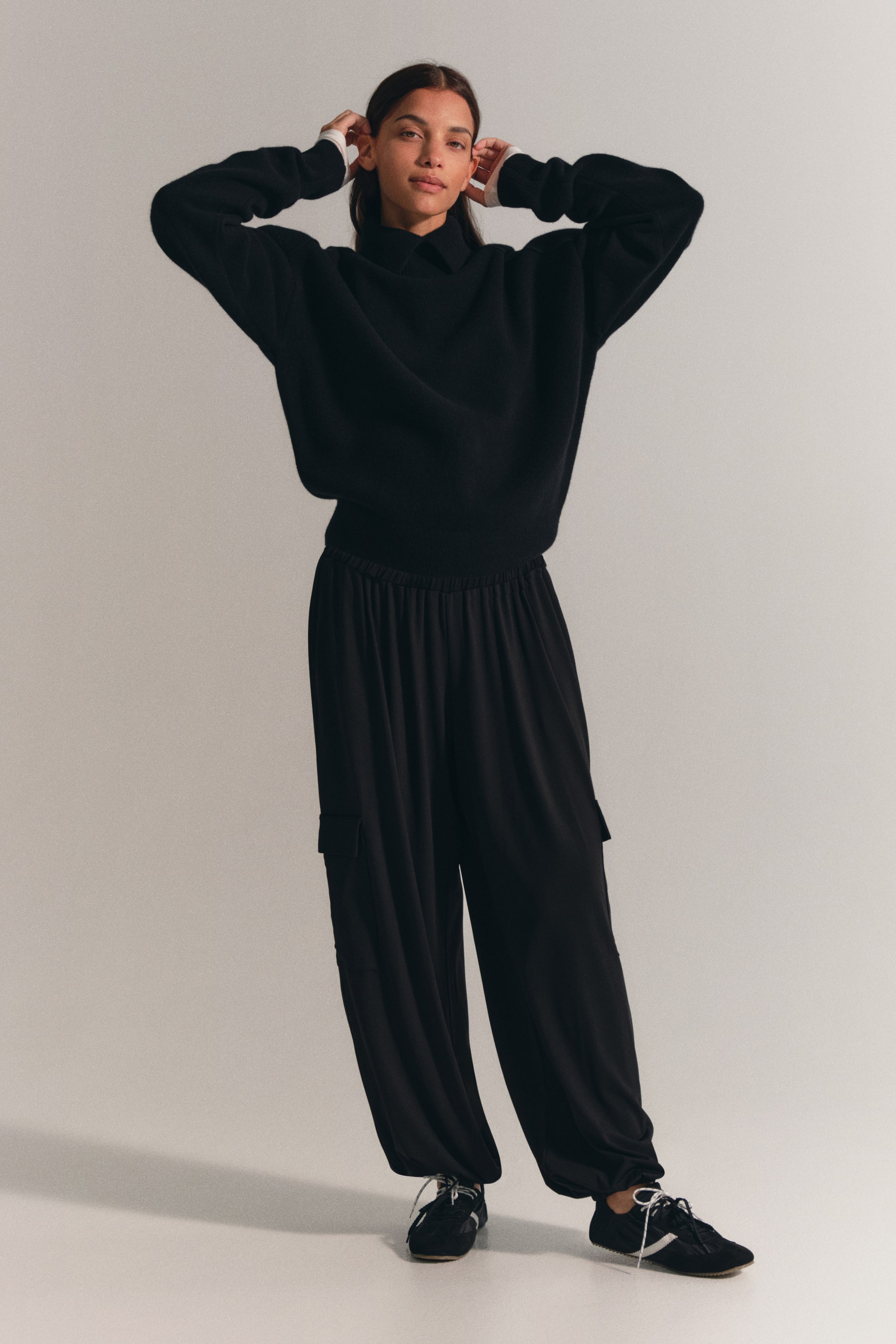 Vezi imaginea mai mare: Pantaloni balloon negri, cu croi pe picior, din jerseu elastic, purtați de model, cu o croială lejeră, talie joasă elastică, buzunare laterale cu clapă și tivuri elastice strânse la glezne. Pantalonii sunt asortați cu un pulover negru cu mânecă lungă și pantofi sport negri cu profil jos.