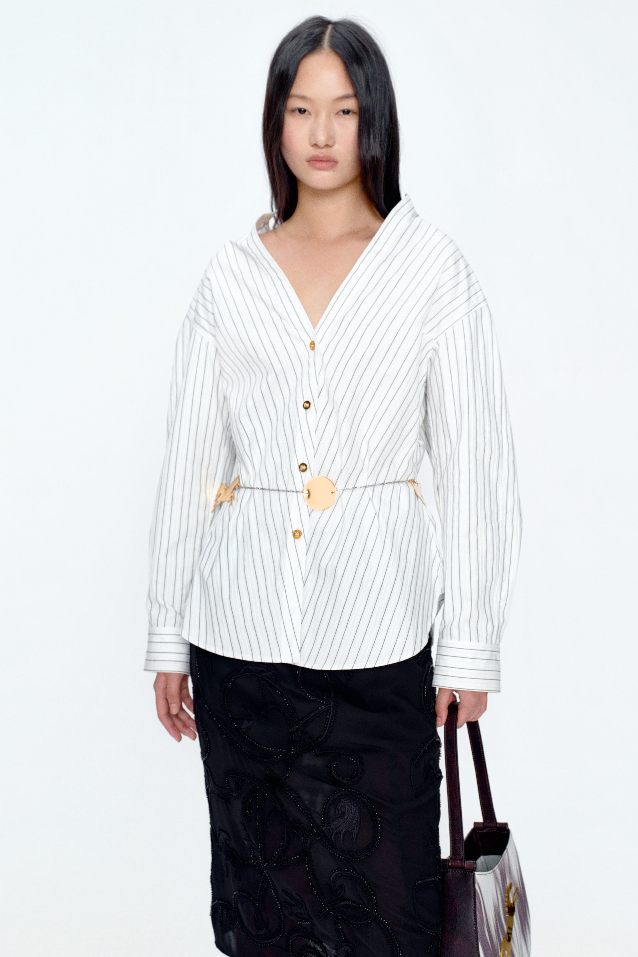 View larger image: A white, long-sleeve shirt with fine vertical black stripes is shown from the front, featuring a V-neckline, open collar and gold-tone buttons along the front. The shirt is cinched at the waist with a slim chain belt detailed with circular light-toned discs, and is styled over a black knee-length skirt with intricate tonal embroidery.