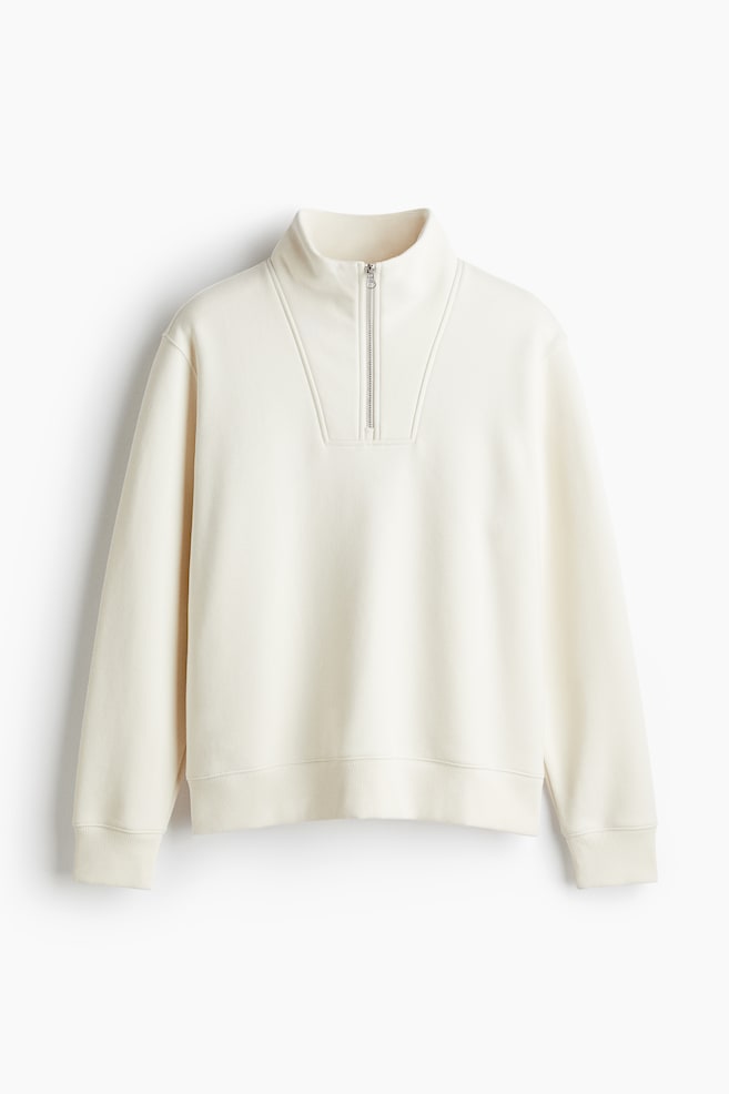 A cream-colored sweatshirt is being presented from the FRONT, featuring a stand-up collar and a silver-toned half-zip closure. It is designed with dropped shoulders, long sleeves with ribbed cuffs, a ribbed hem, and two stitched lines are forming a V-shape on the chest. - 1